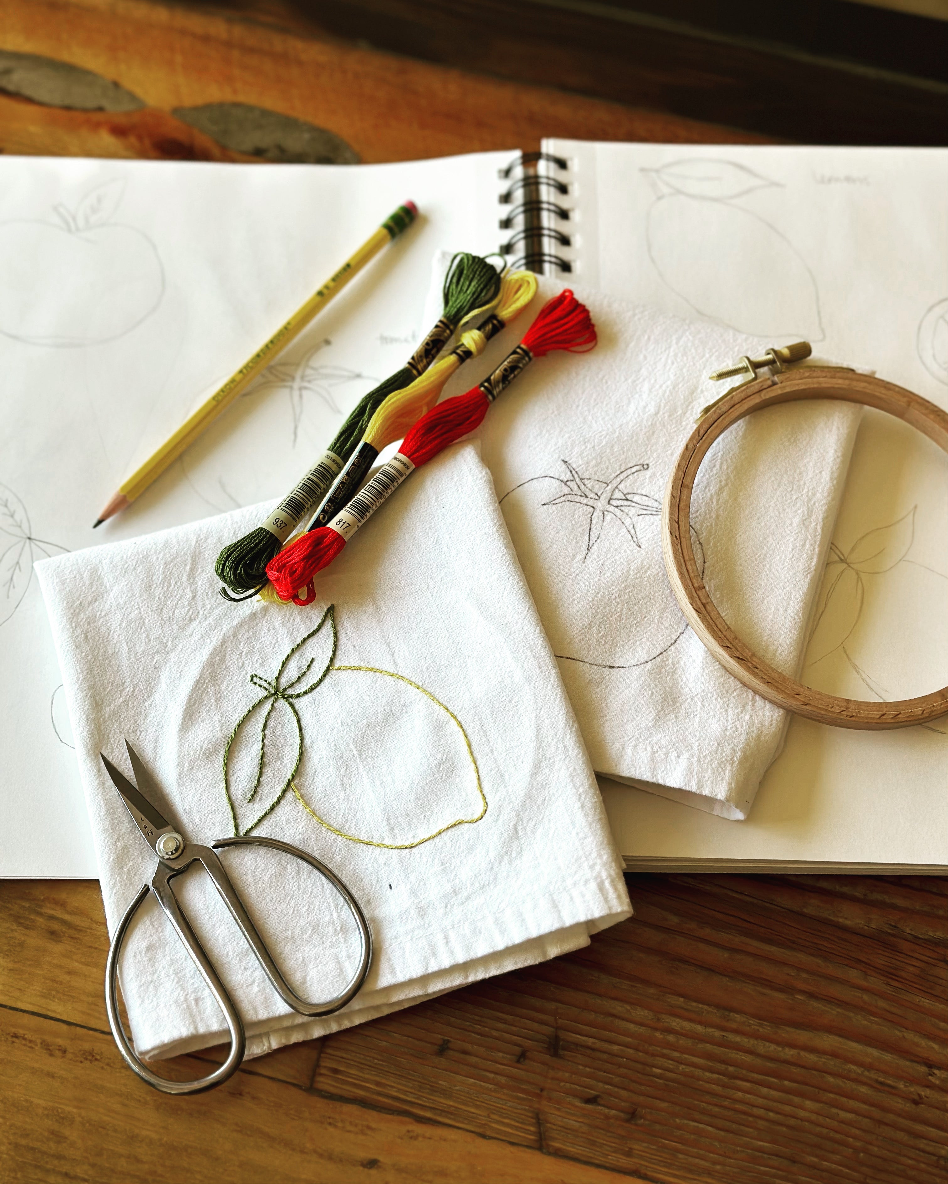 Embroidery materials including scissors, thread, and an embroidery hoop on an open sketchbook with pencil on a wooden surface.