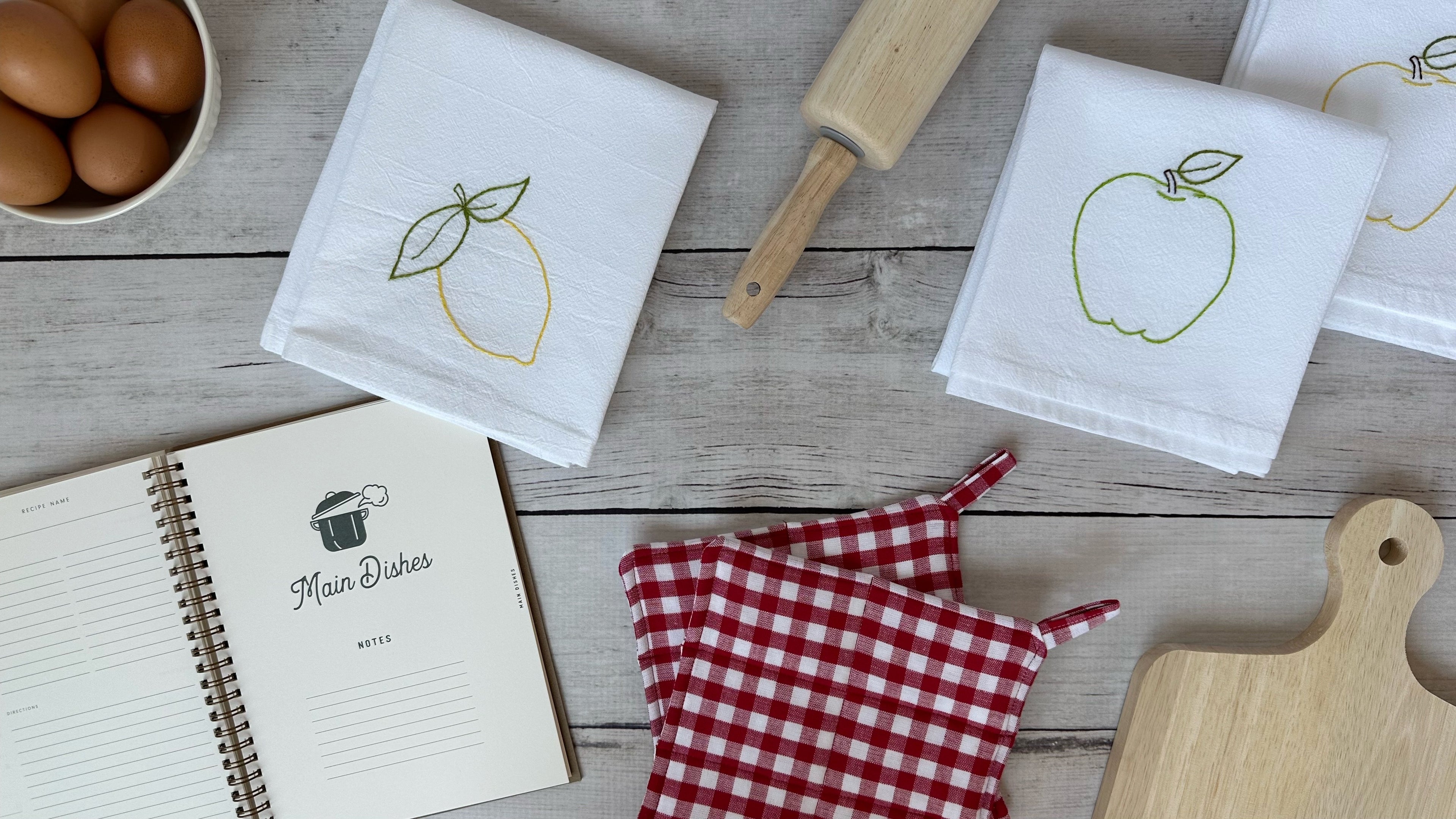 Kitchen-themed items including tea towels with fruit designs, hot pads, a notebook, a rolling pin, eggs, and a cutting board on a wooden surface.