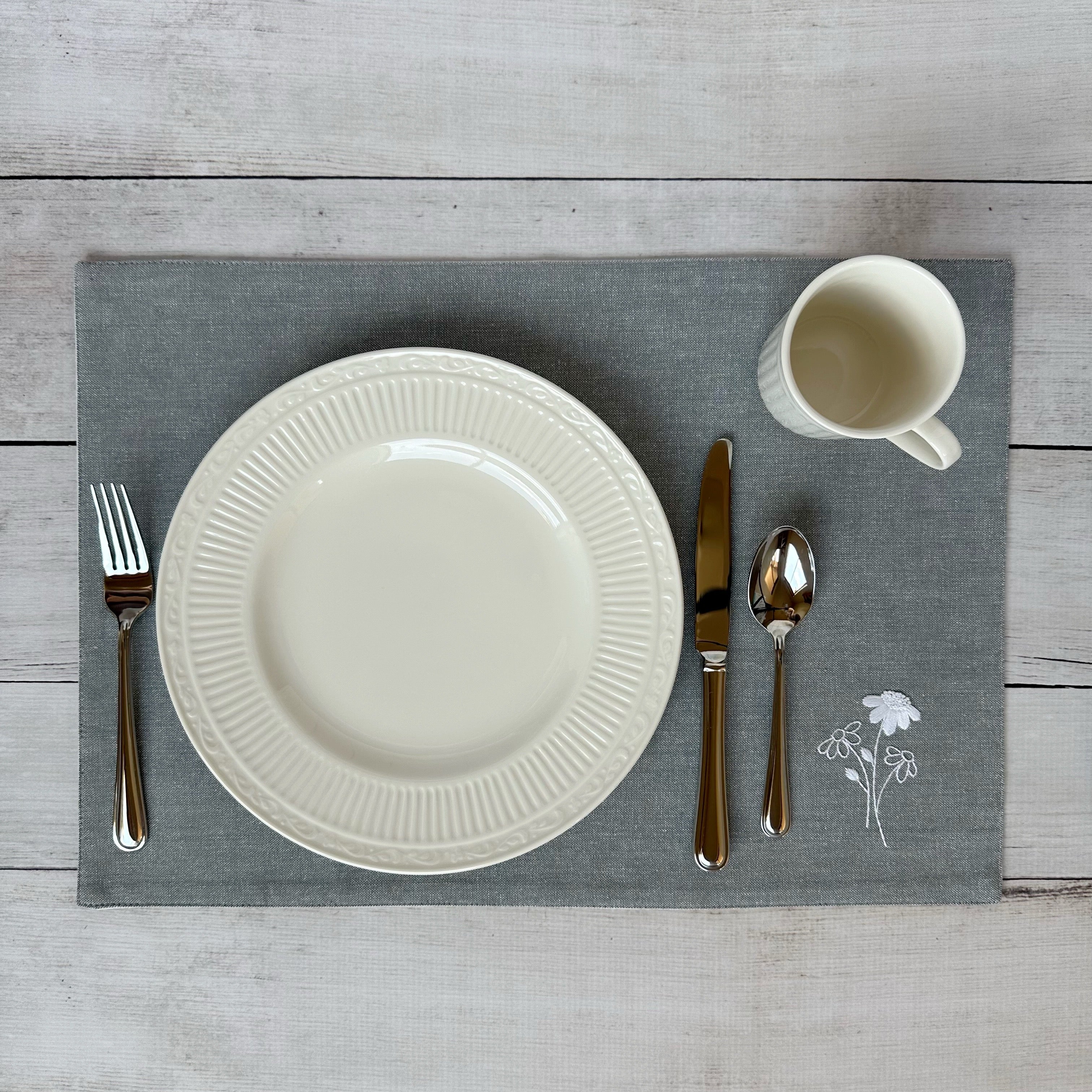 White china plate with silverware on a gray placemat with embroidered flowers on a wooden surface