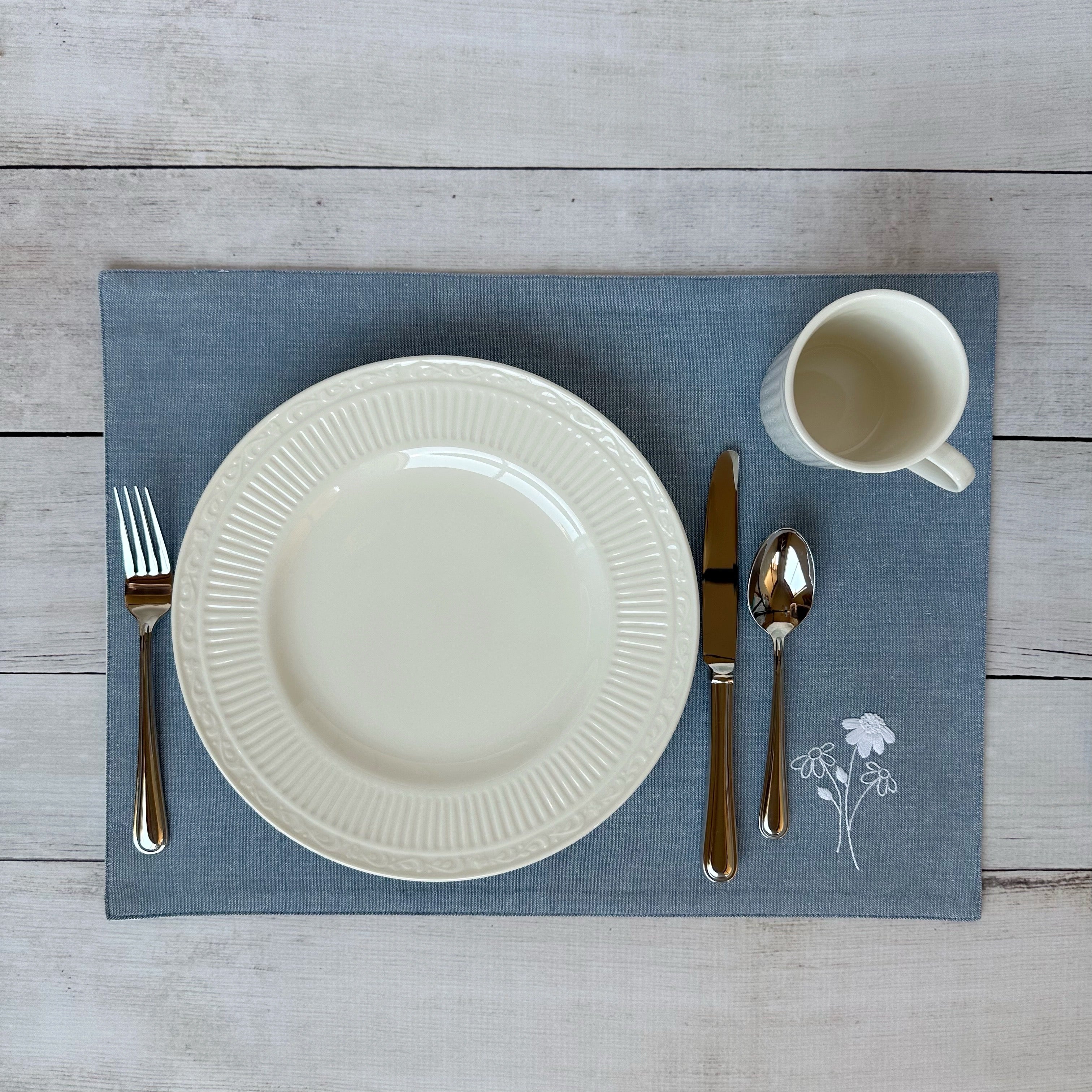 White china plate with silverware on a blue placemat with embroidered flowers on a wooden surface