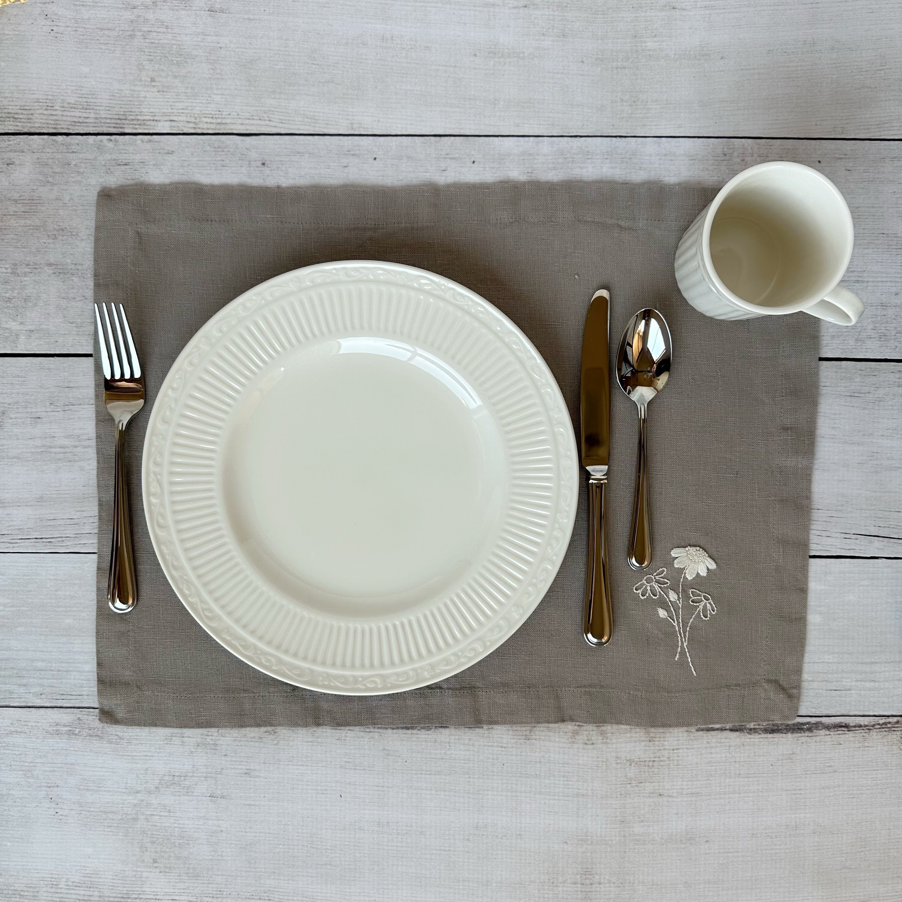 White china plate with silverware on a taupe placemat with embroidered flowers on a wooden surface