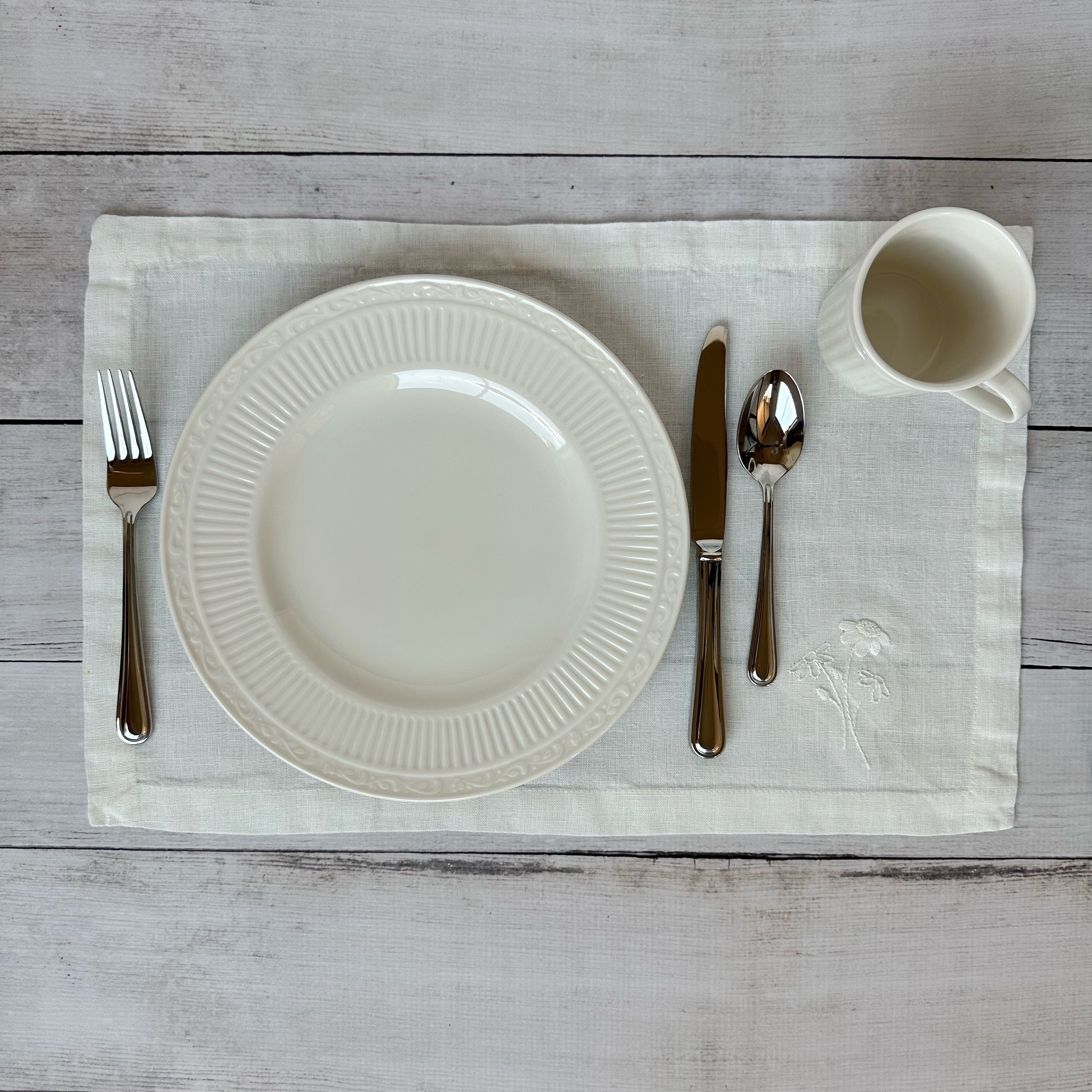 White china plate with silverware on an ivory placemat with embroidered flowers on a wooden surface