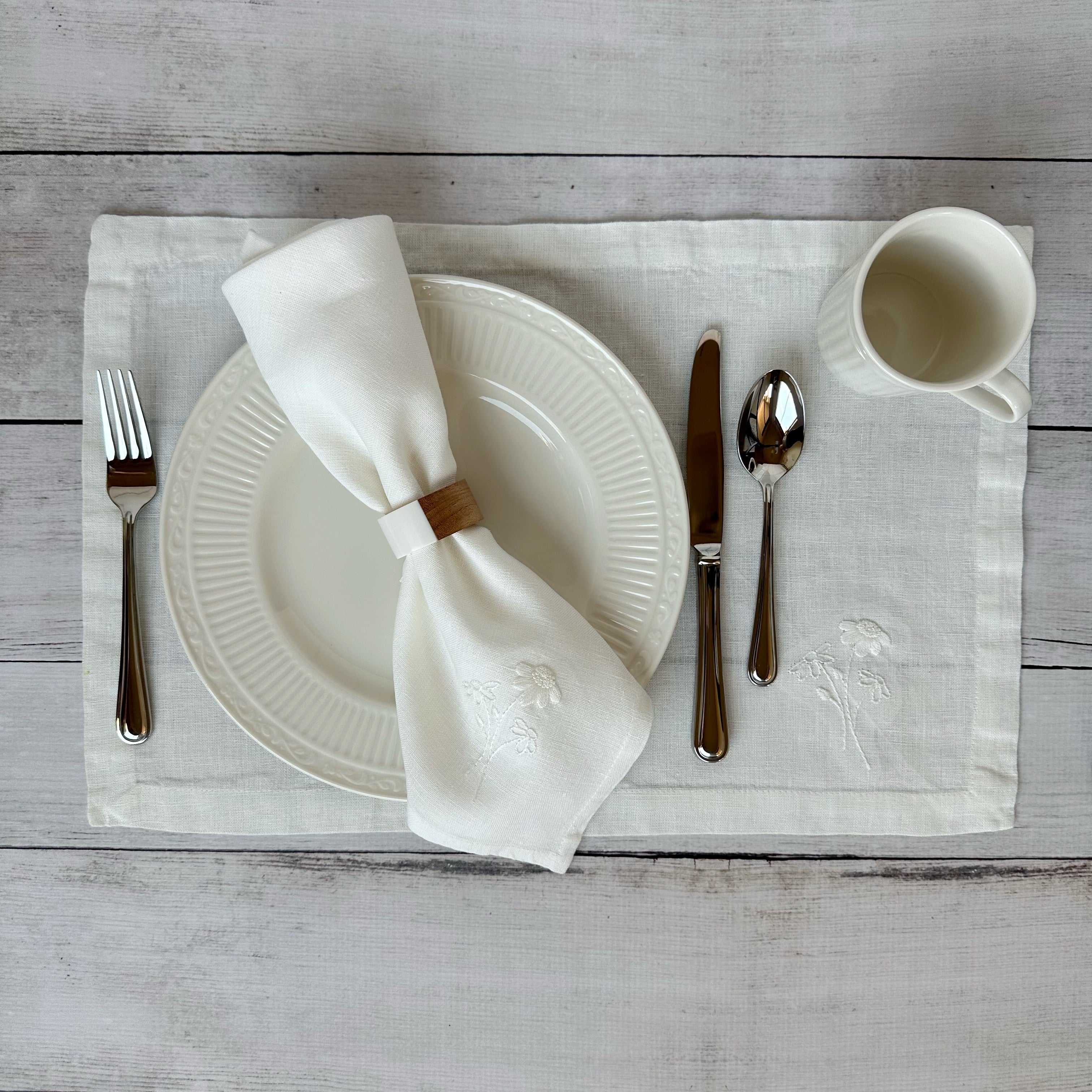 Dining table setting with white plates, silverware, and embroidered linen napkin and placemat on a wooden surface.