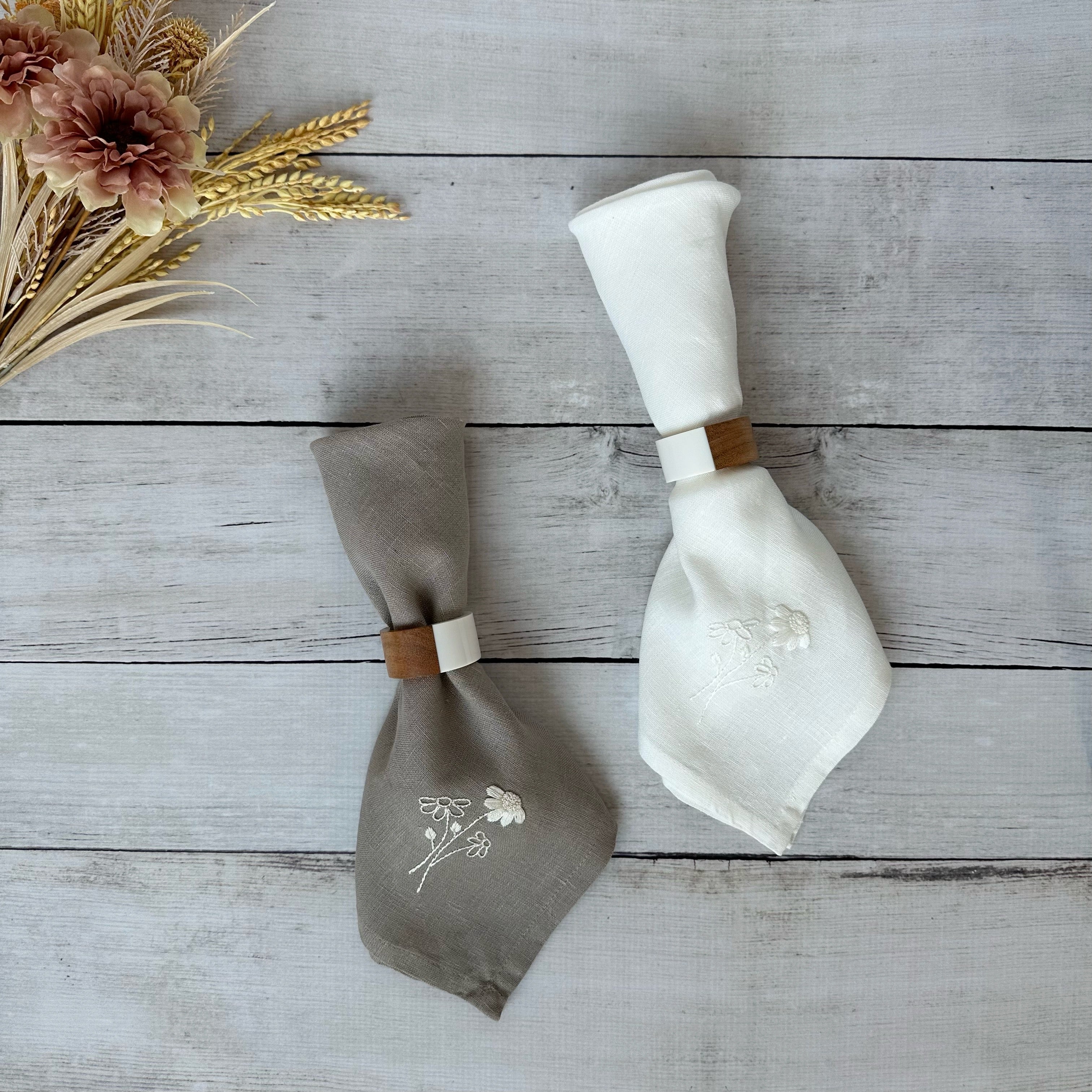 Taupe and ivory linen napkins with embroidered flower design with wooden napkin rings on a wooden surface