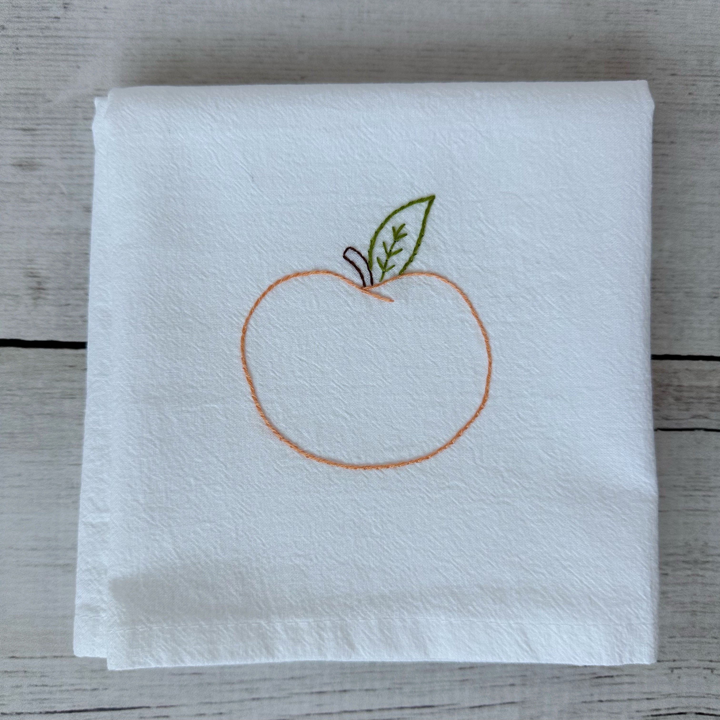 White tea towel with embroidered peach design on a wooden surface