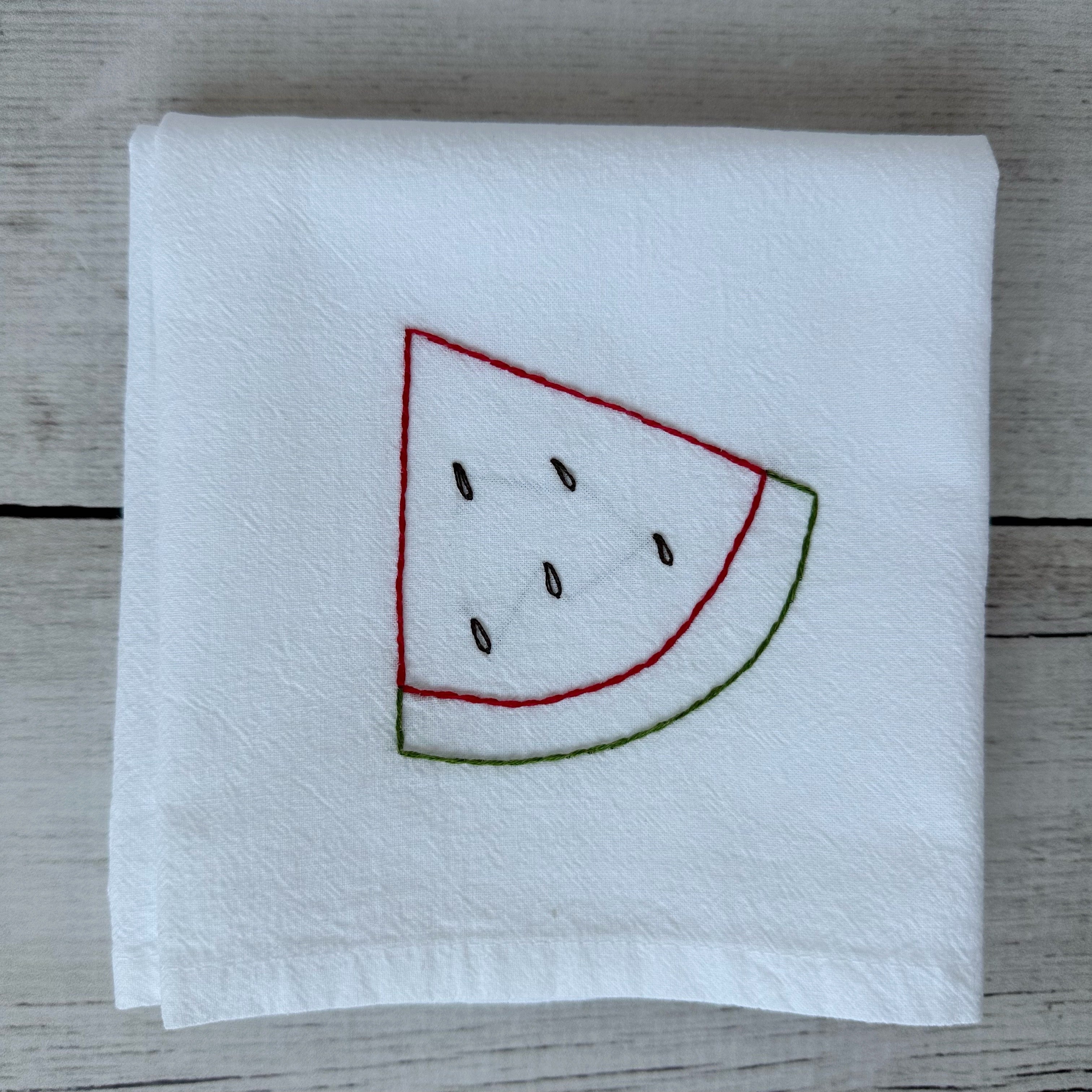 White tea towel with embroidered watermelon slice design on a wooden surface