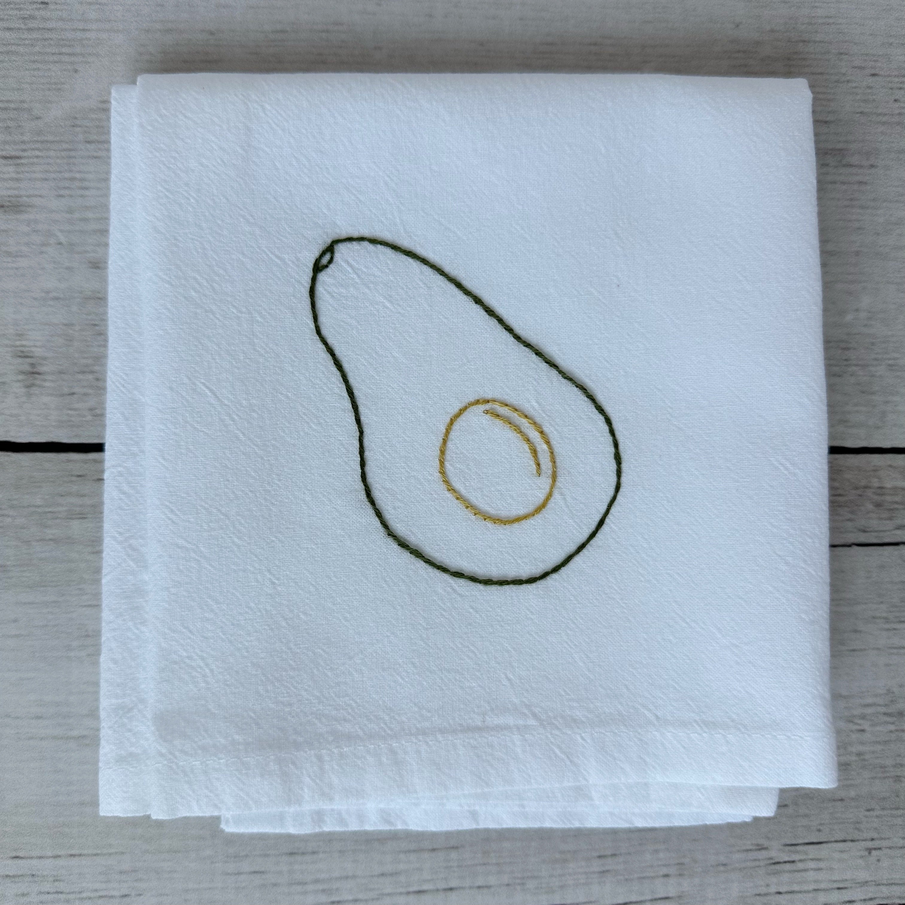 White tea towel with embroidered avocado design on a wooden surface
