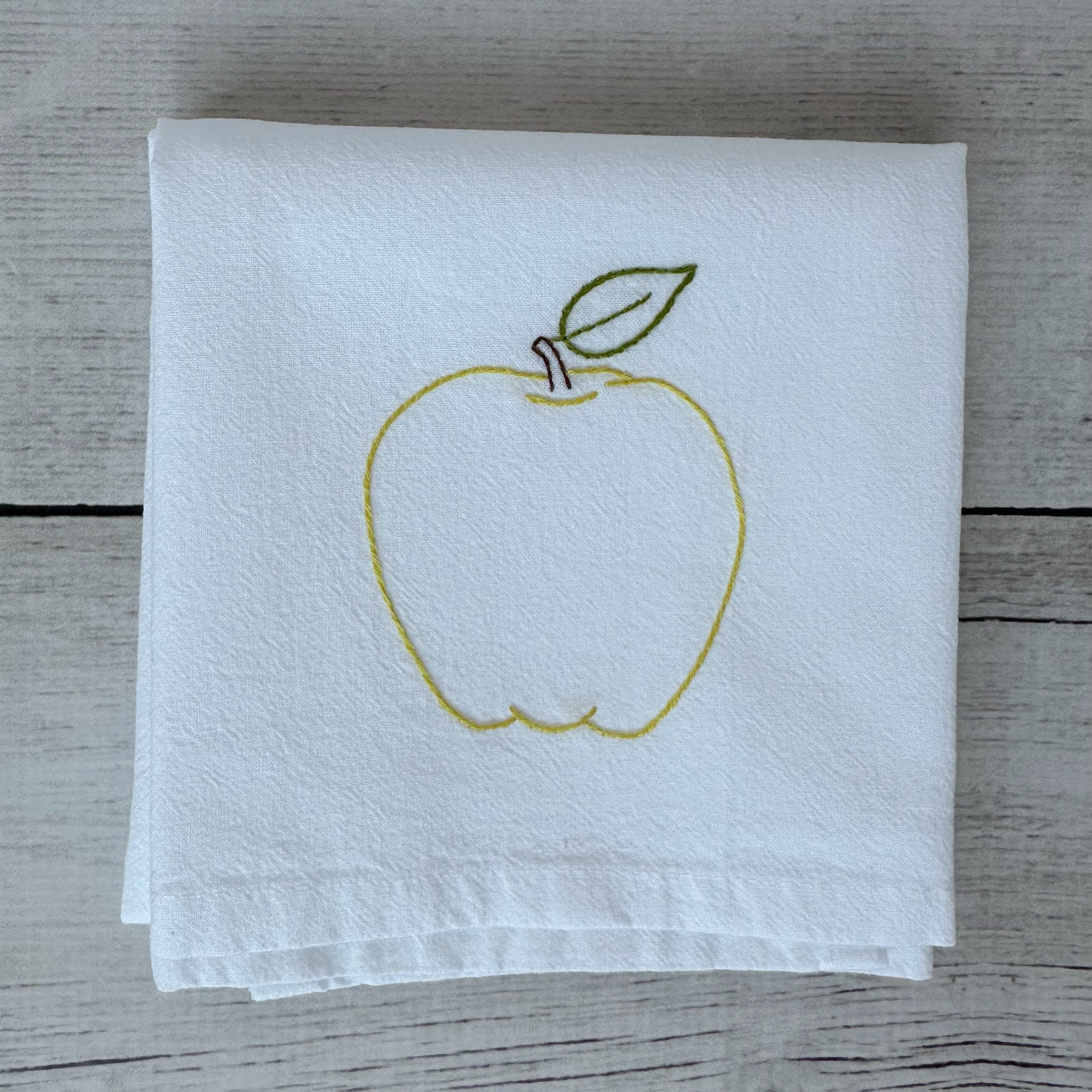 White tea towel with embroidered yellow apple design on a wooden surface