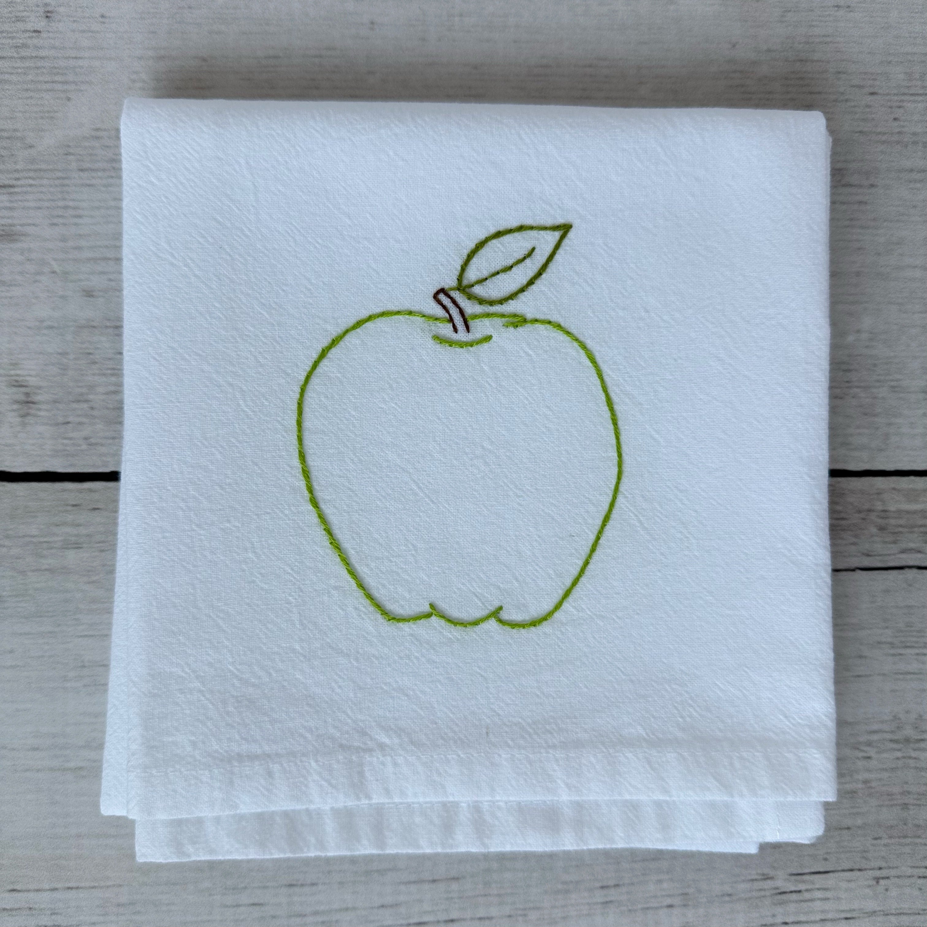 White tea towel with a green embroidered apple design on a wooden surface