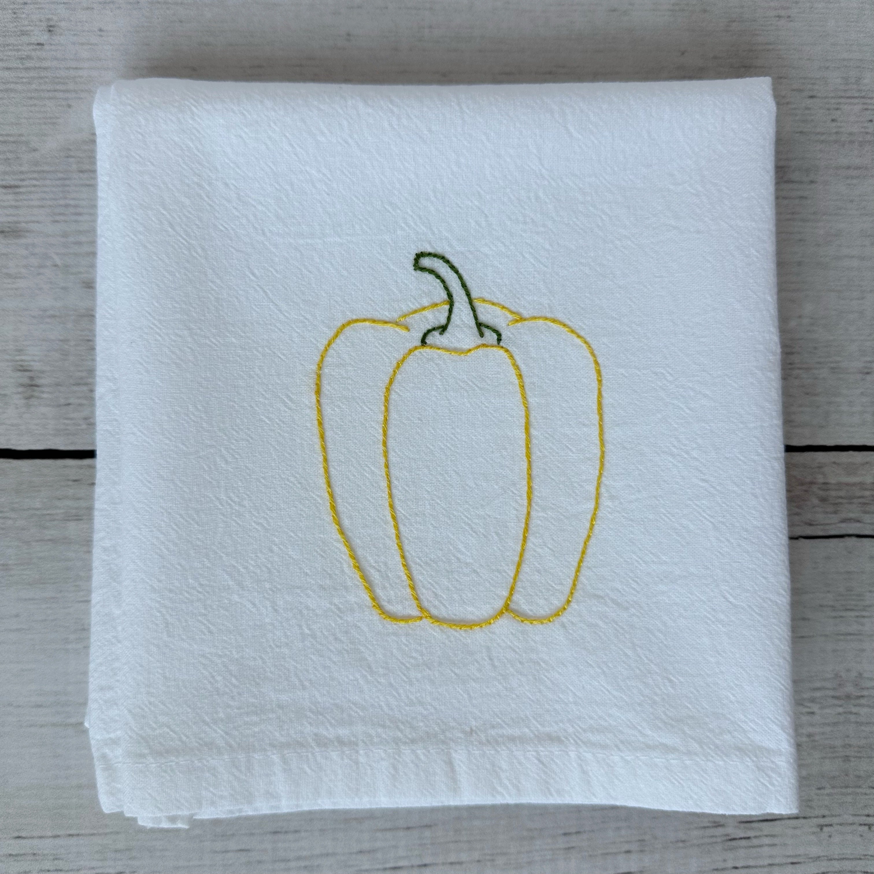 White tea towel with embroidered yellow bell pepper design on a wooden surface