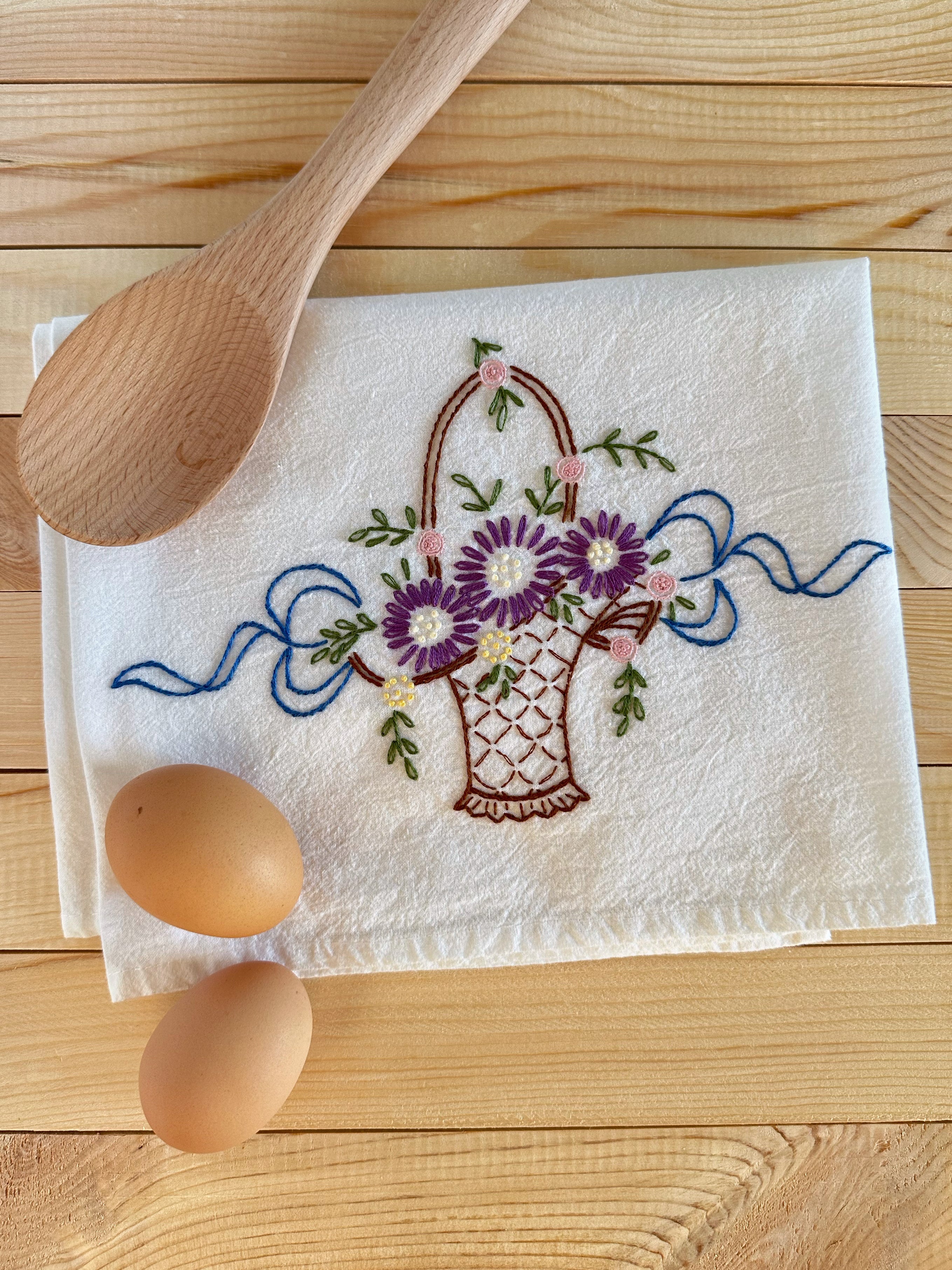 Embroidered towel with floral design on a wooden surface with eggs and a wooden spoon.