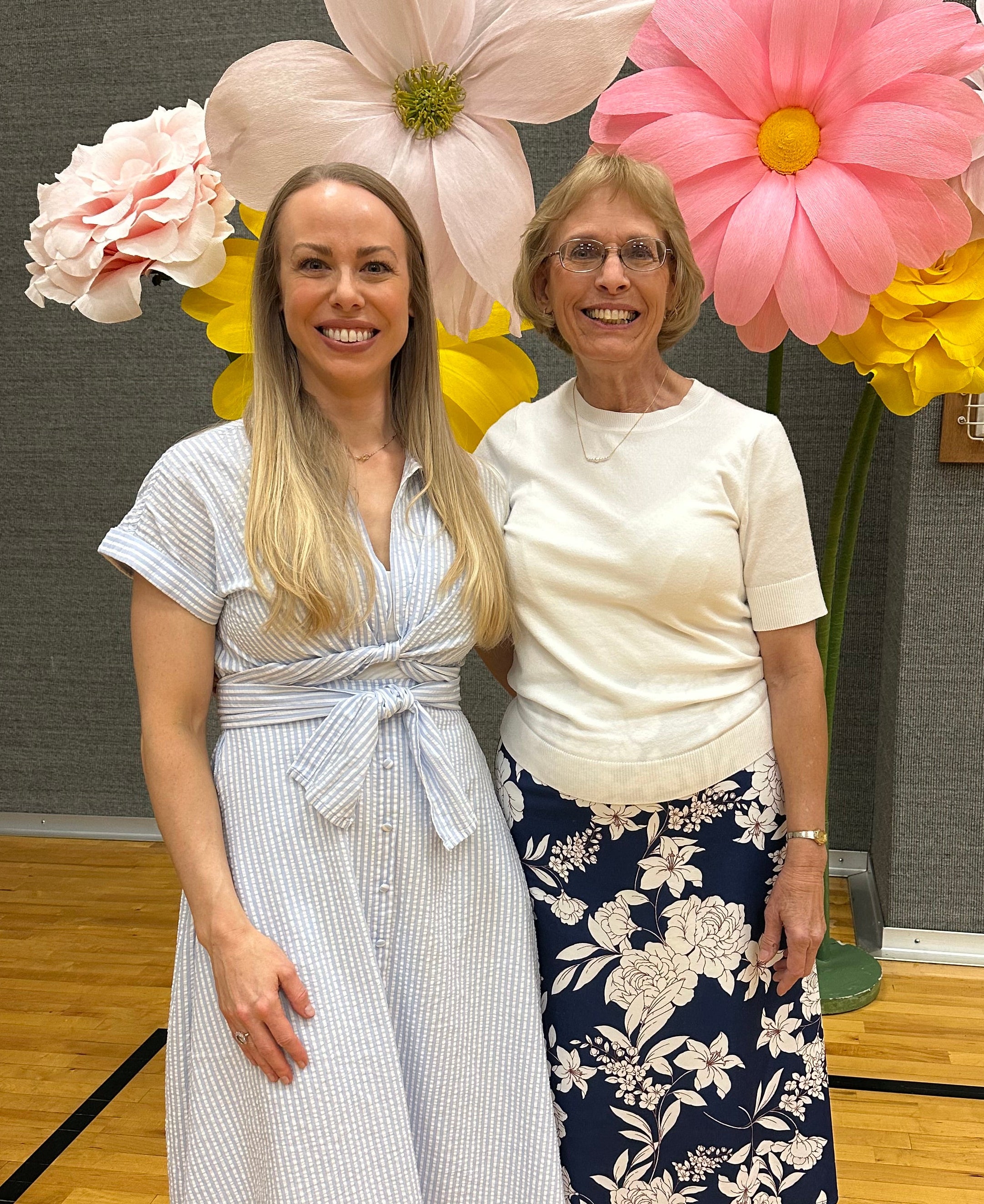 Two women standing next to each other with decorative flowers on a wooden floor.