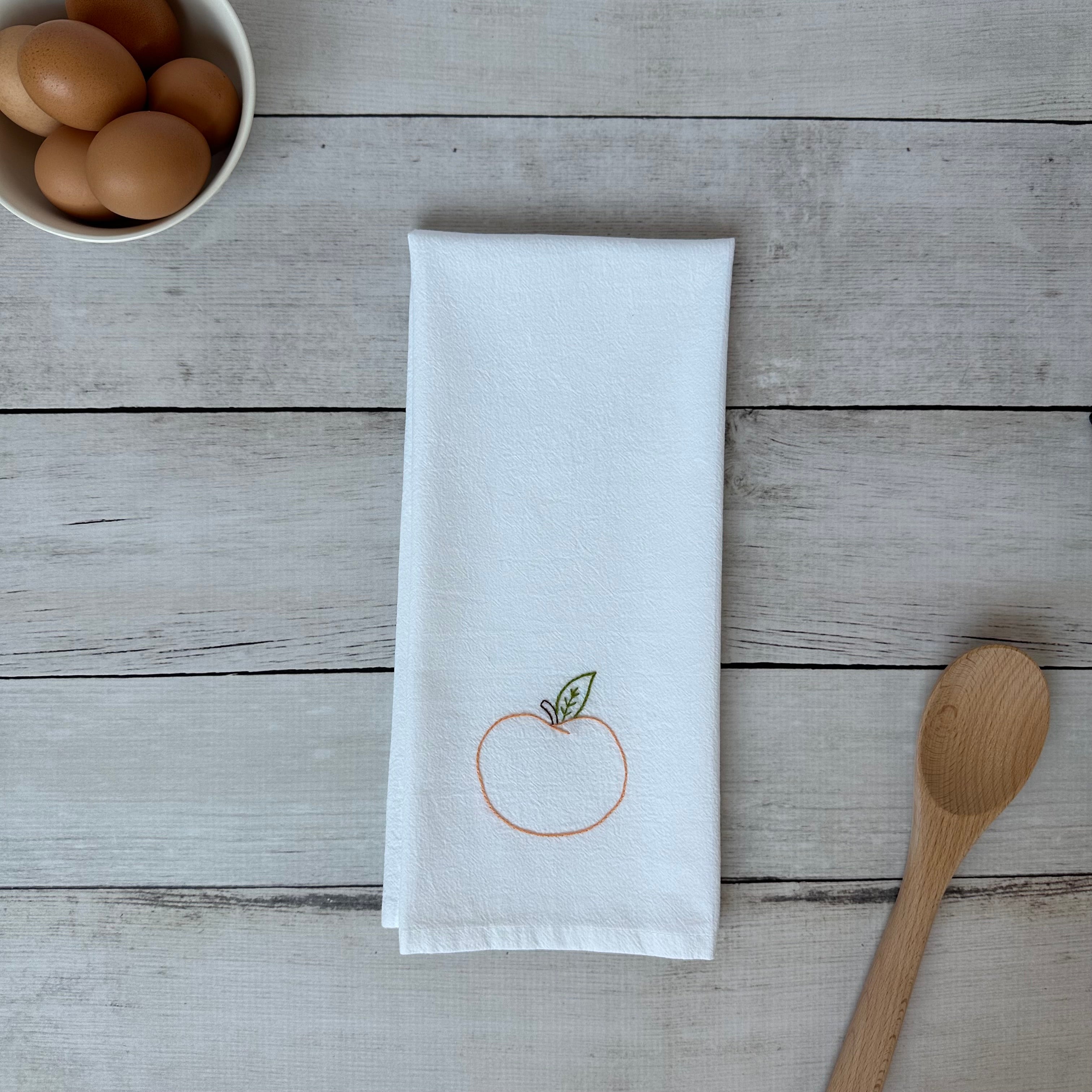White tea towel with embroidered peach design on a wooden surface with eggs and a spoon.
