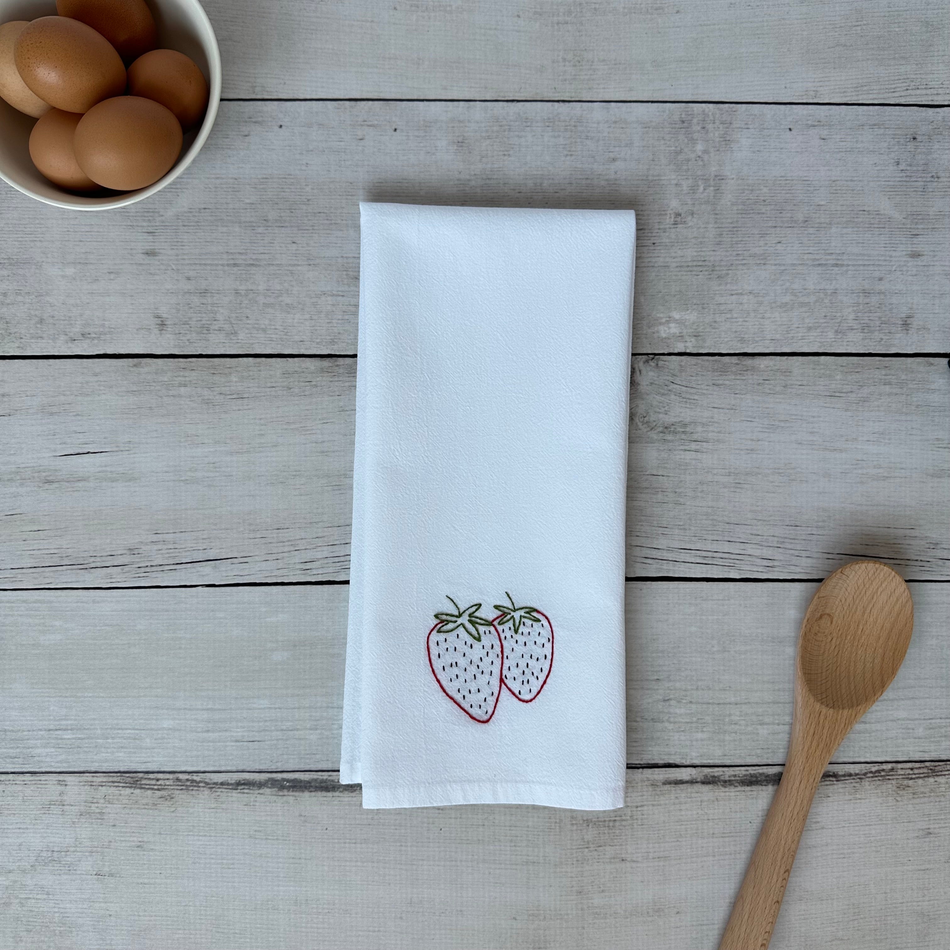 White tea towel with embroidered strawberry design on a wooden surface with eggs and a wooden spoon.