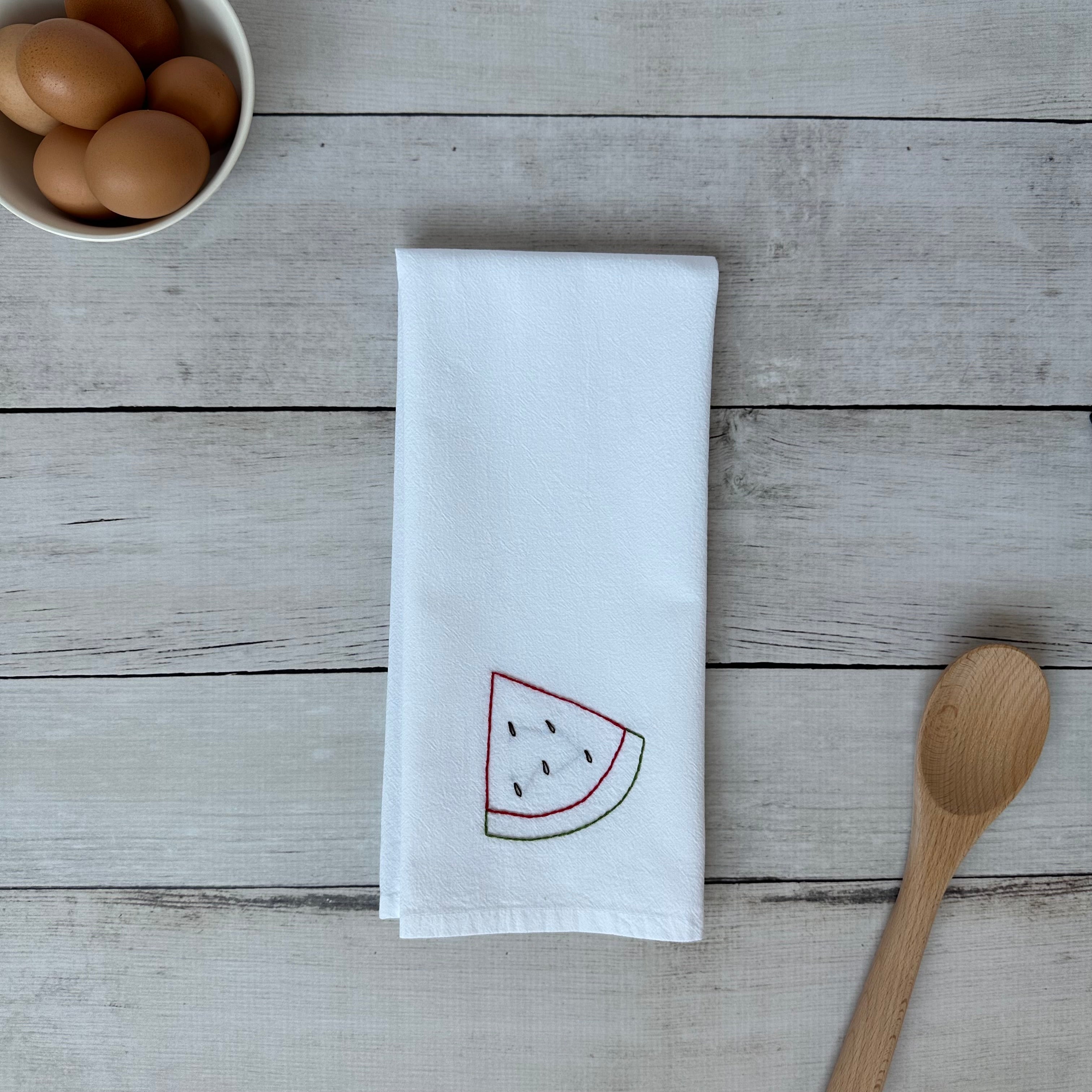 White tea towel with embroidered watermelon design on a wooden surface with eggs and a spoon.