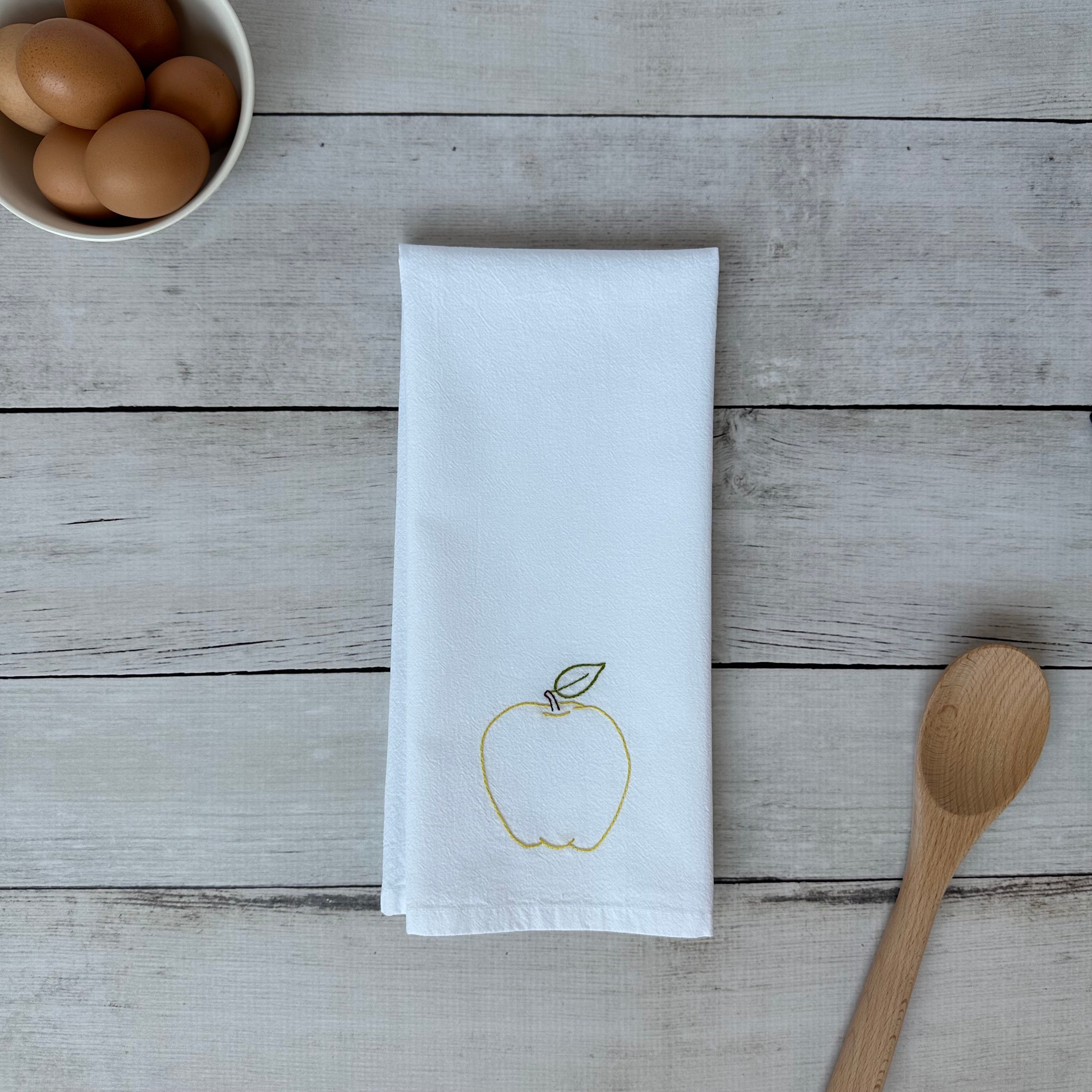 White tea towel with an embroidered yellow apple design on a wooden surface with eggs and a spoon