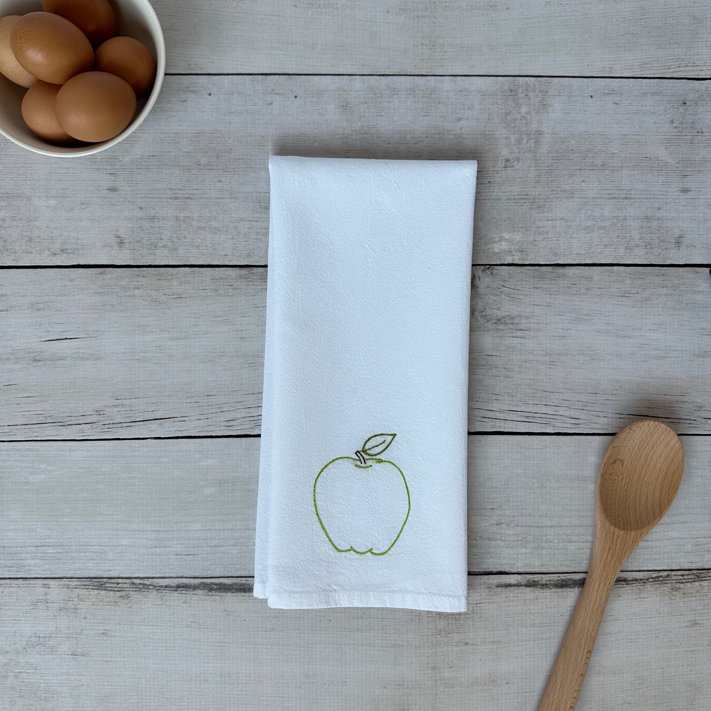 White tea towel with embroidered green apple design on a wooden surface with eggs and a wooden spoon.