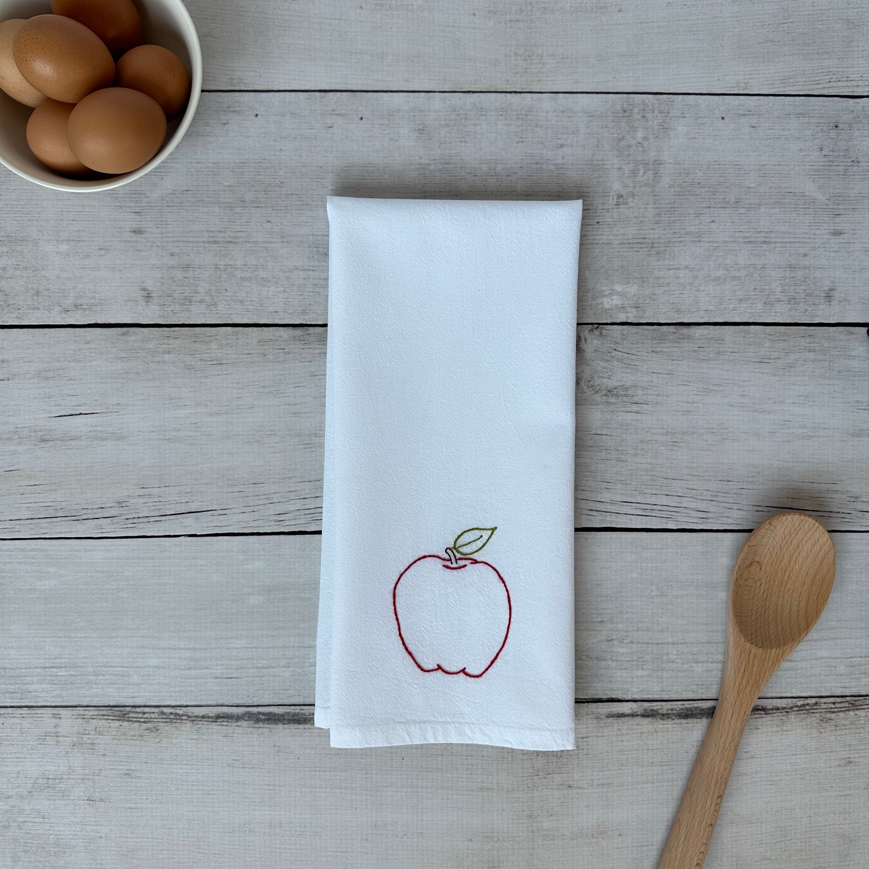 White tea towel with an embroidered red apple design on a wooden surface with eggs and a wooden spoon.
