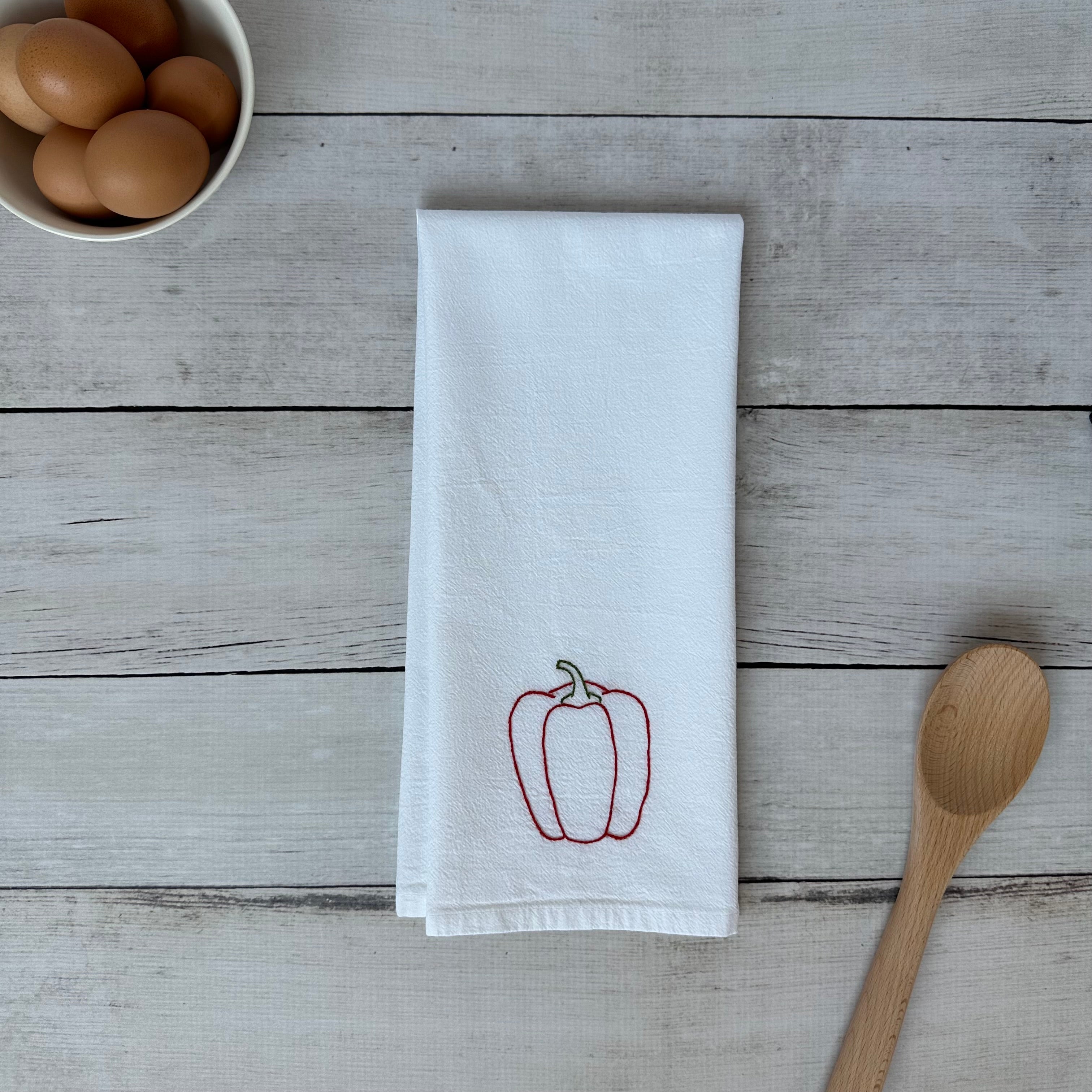 White tea towel with embroidered red bell pepper design on a wooden surface with eggs and a spoon.