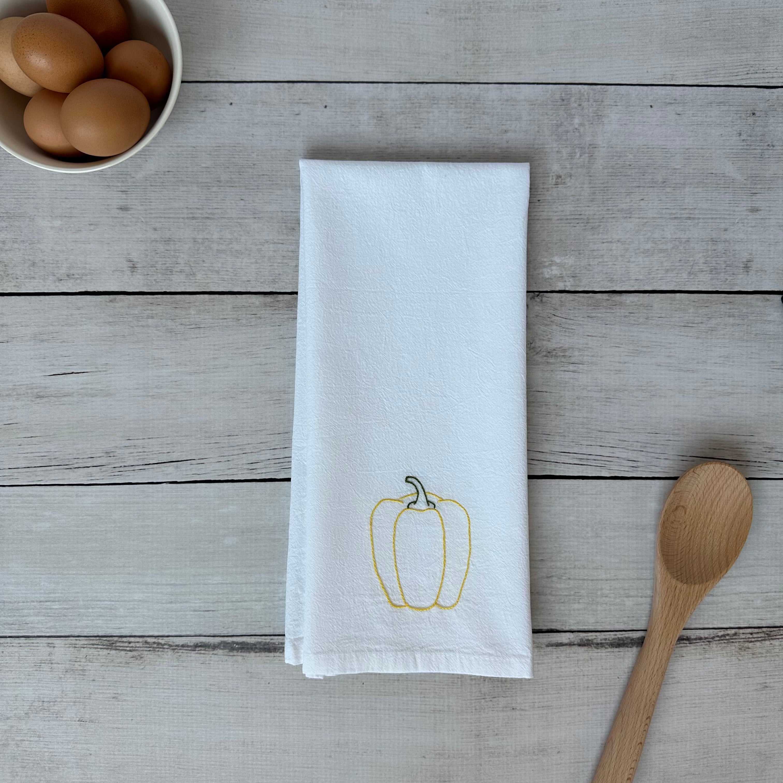 White tea towel with embroidered yellow bell pepper design on a wooden surface with eggs and a spoon.