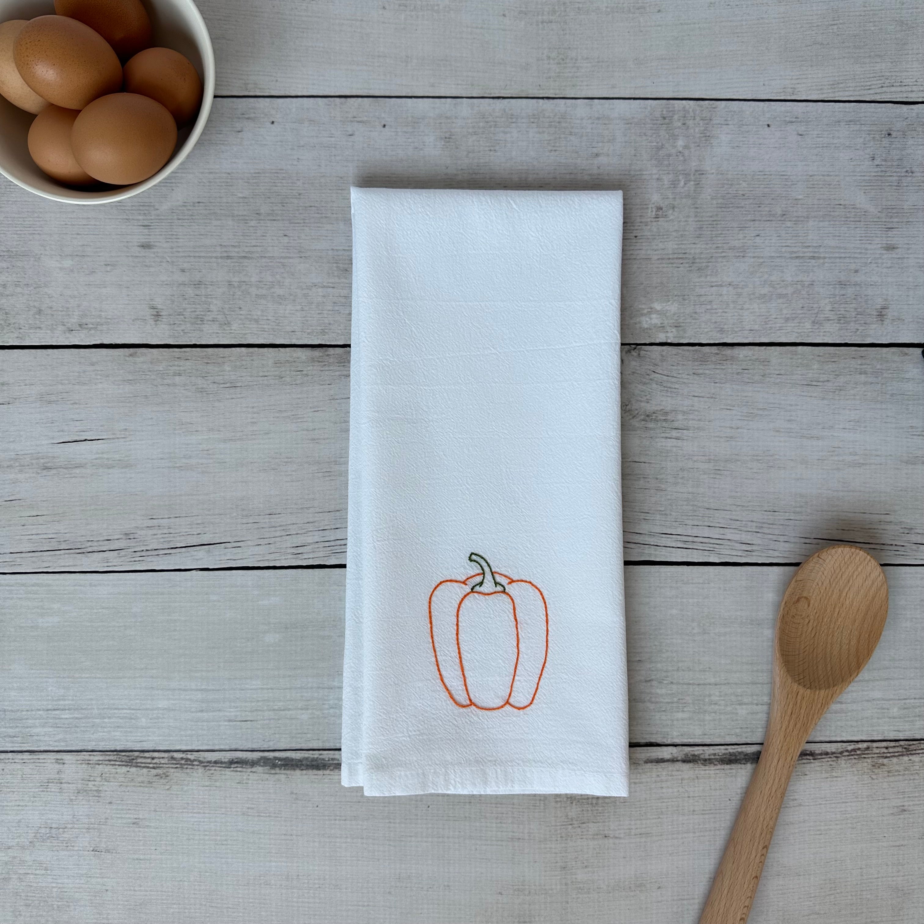 White tea towel with embroidered orange bell pepper design on a wooden surface with eggs and a spoon.