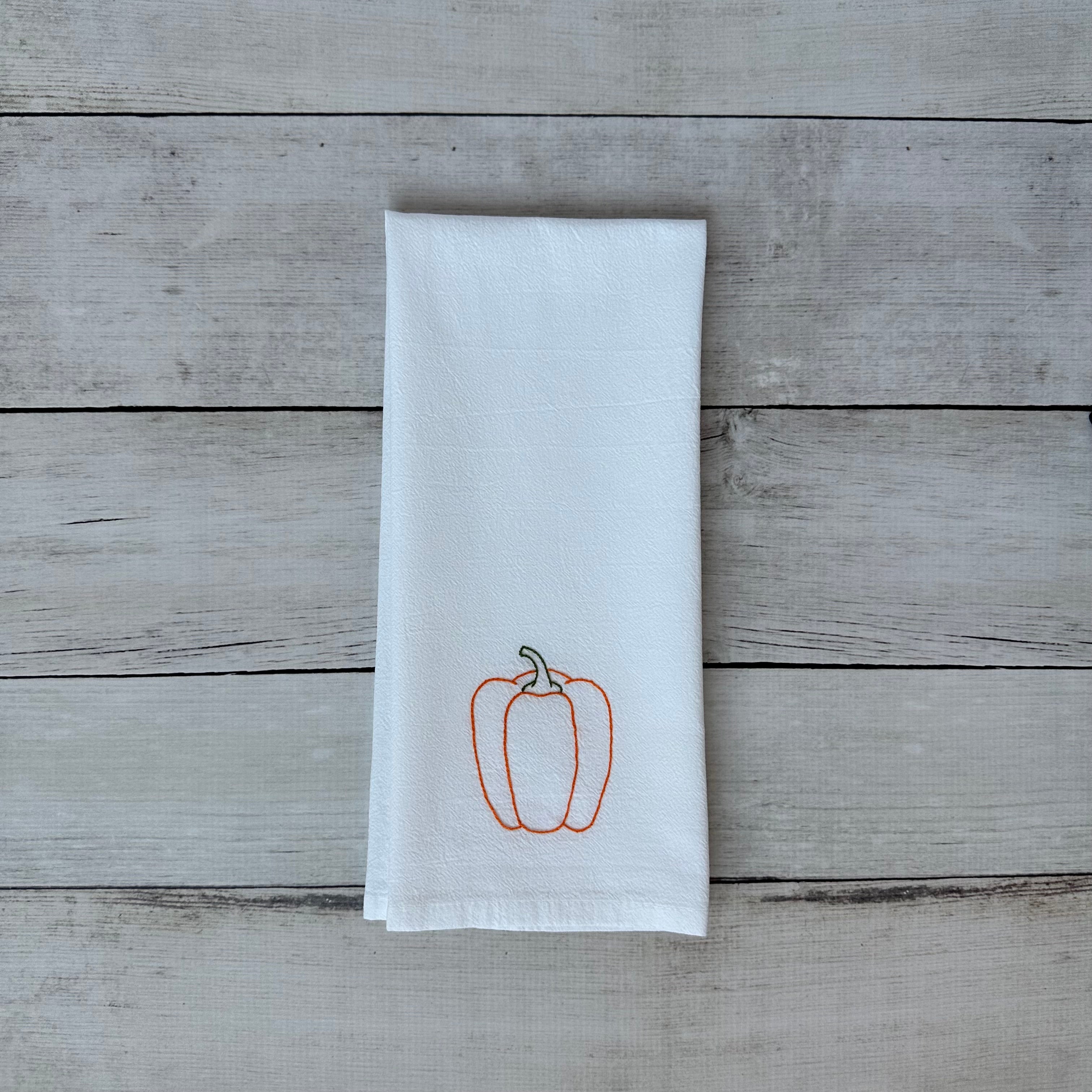 White tea towel with embroidered orange bell pepper design on a wooden surface