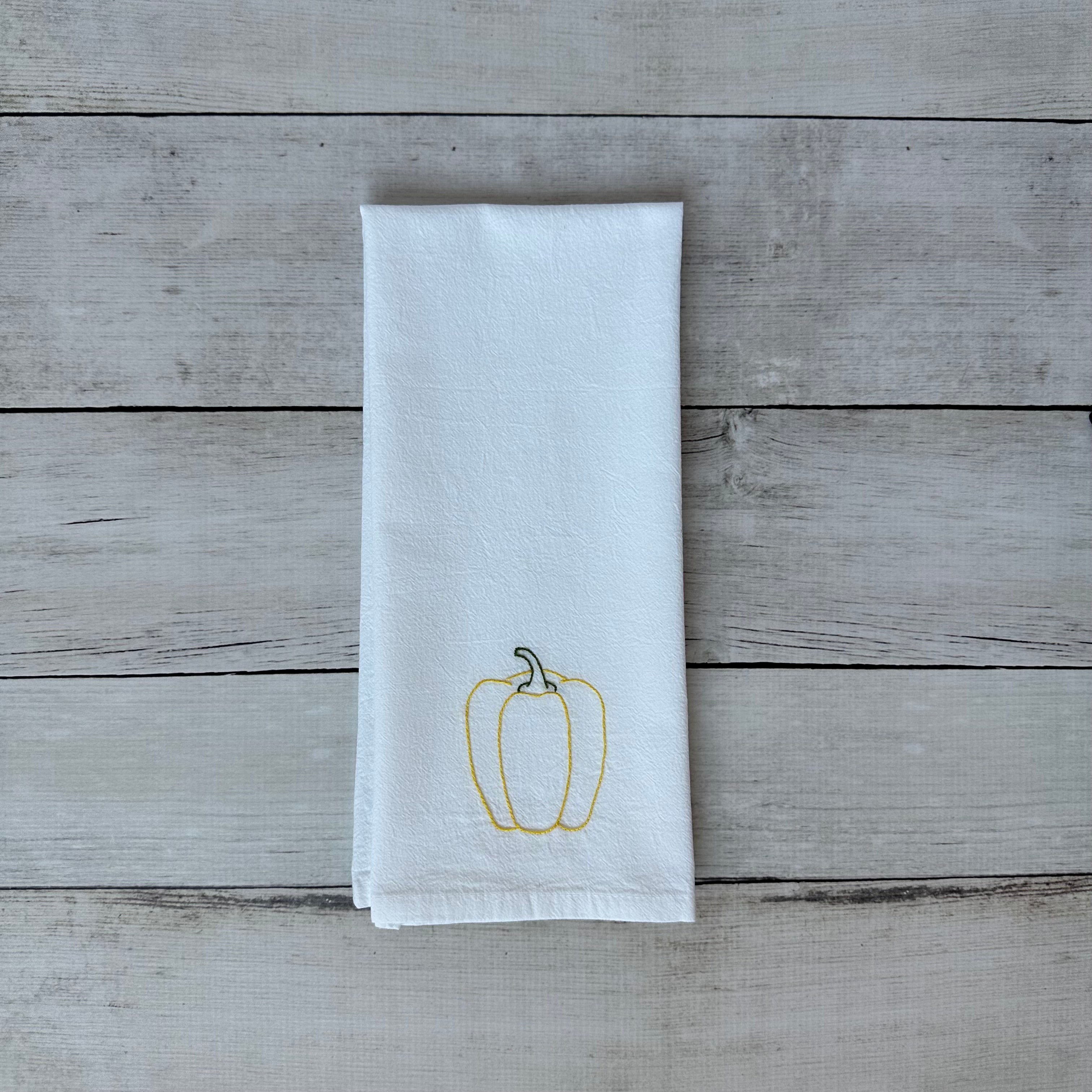White tea towel with embroidered yellow bell pepper design on a wooden surface