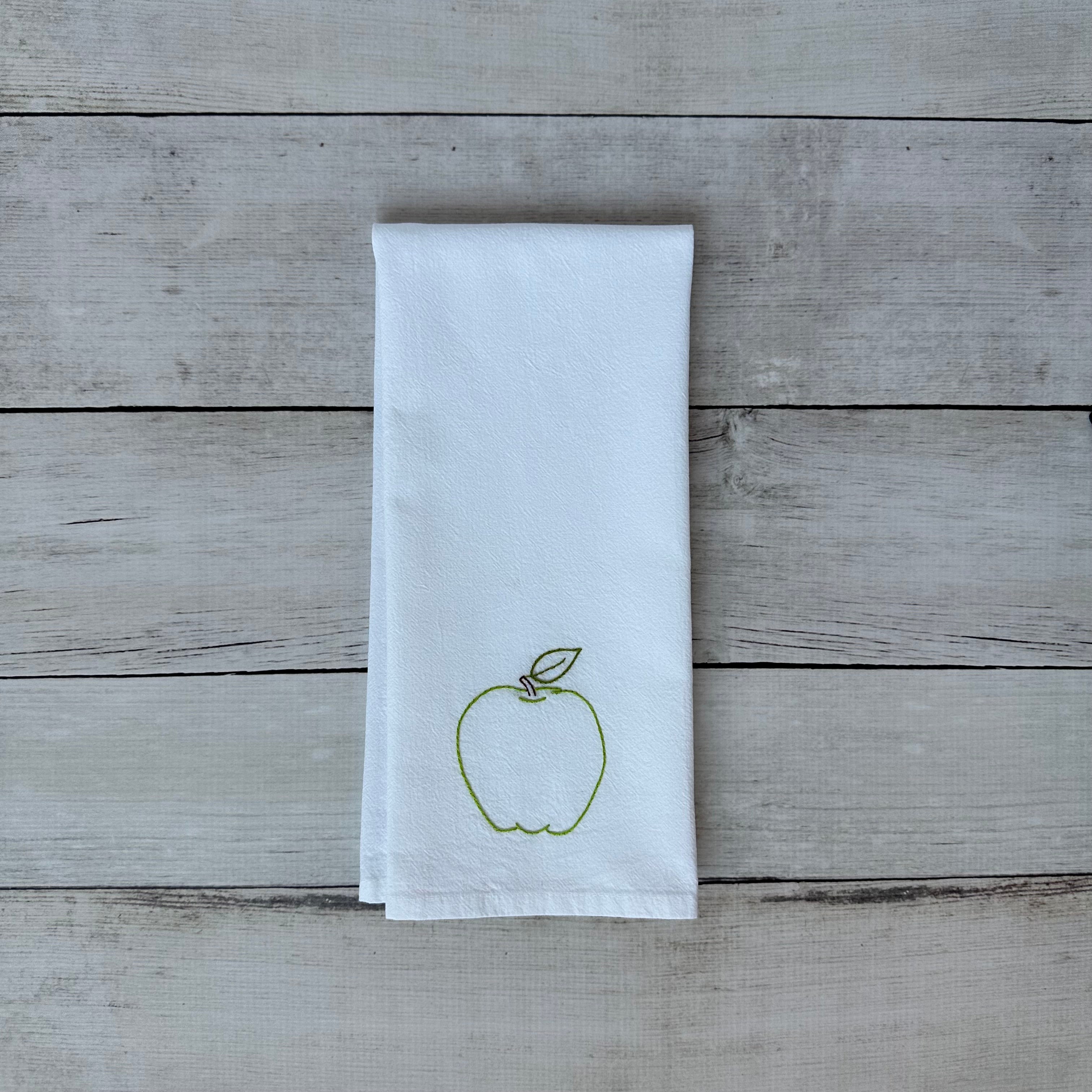 White tea towel with embroidered green apple design on a wooden surface