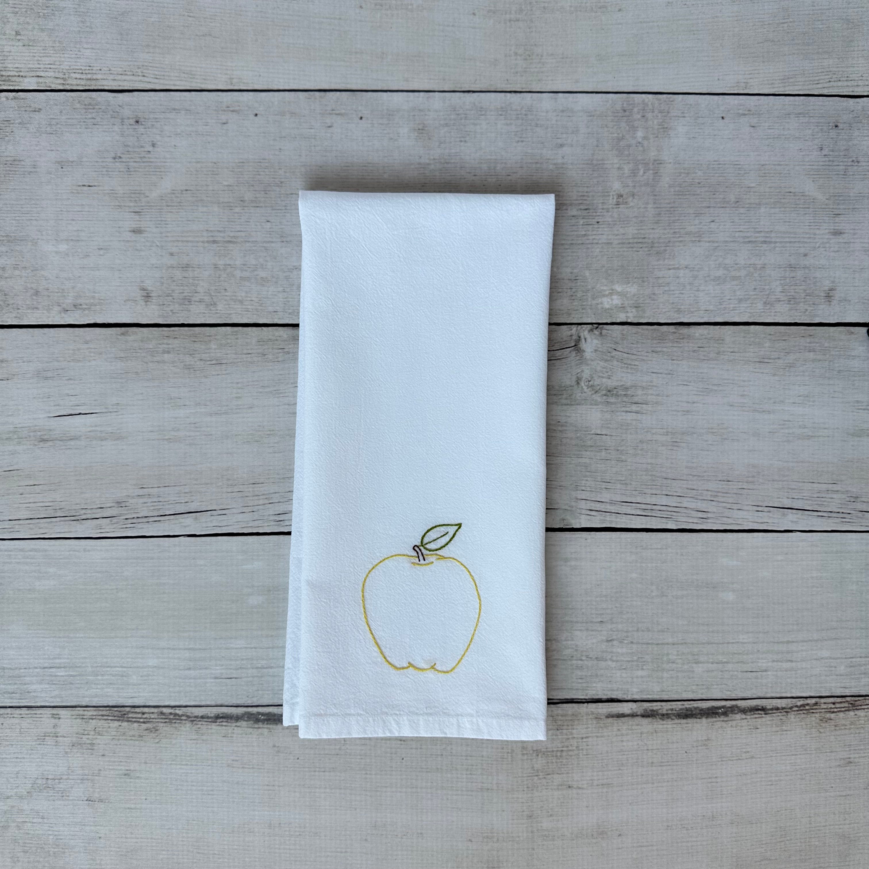 White tea towel with an embroidered yellow apple design on a wooden surface