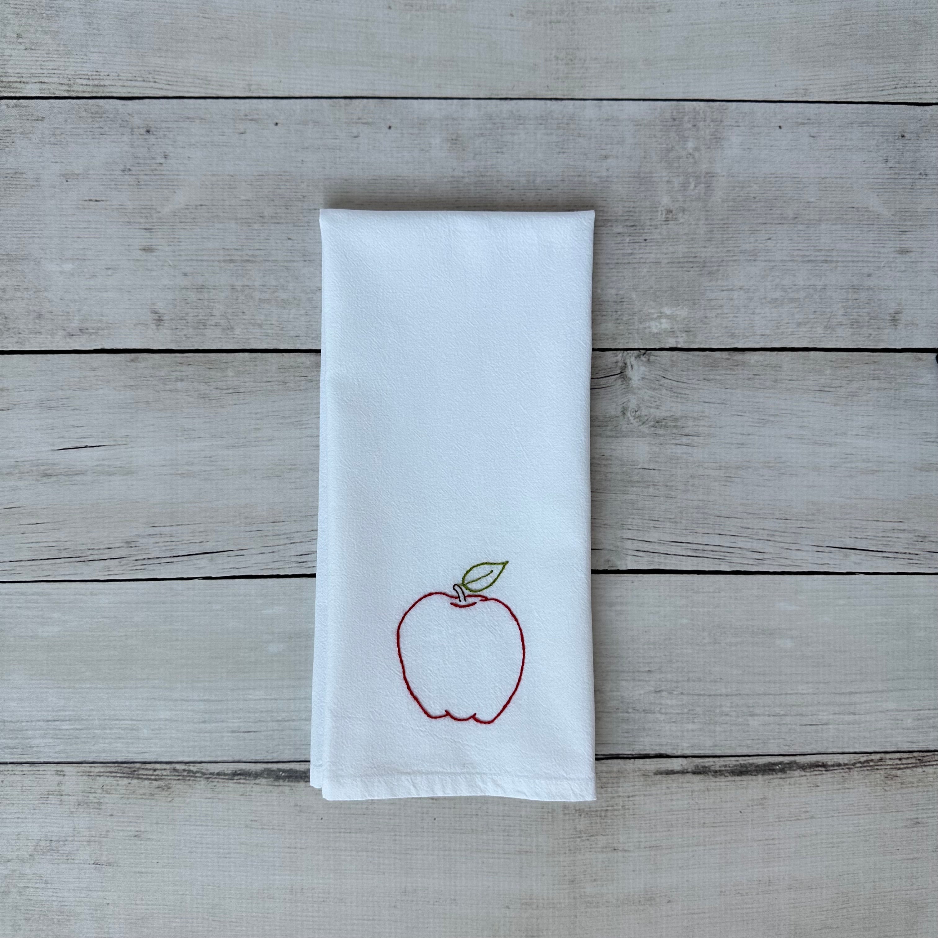White tea towel with an embroidered red apple design on a wooden surface