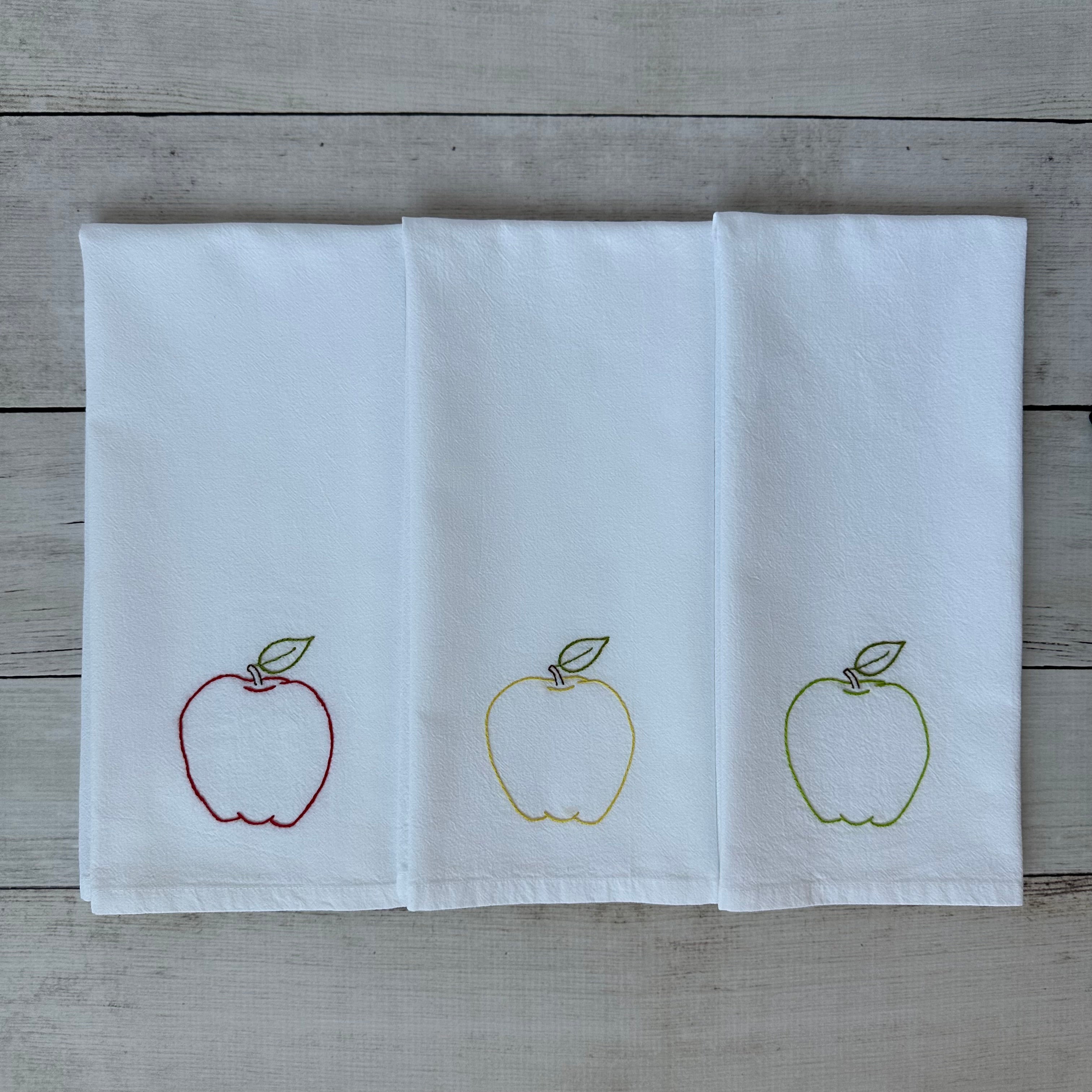 Three white tea towels with embroidered apple designs on a wooden surface