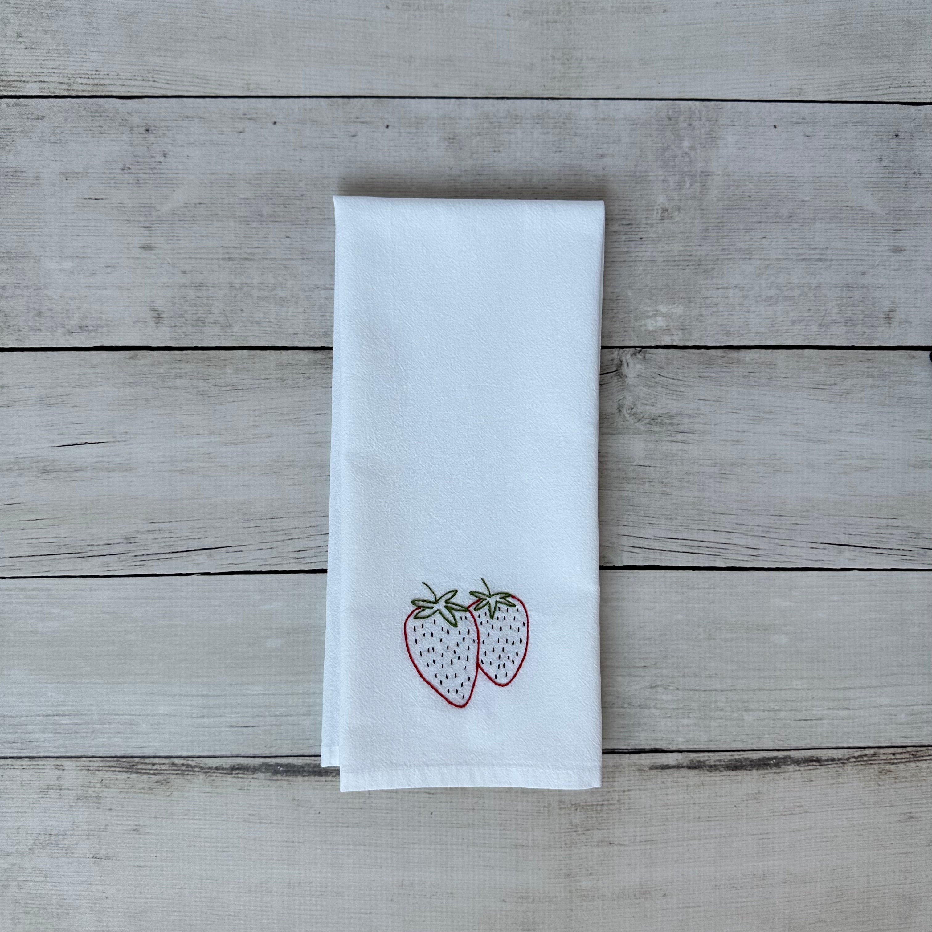 White tea towel with embroidered strawberry design on a wooden surface