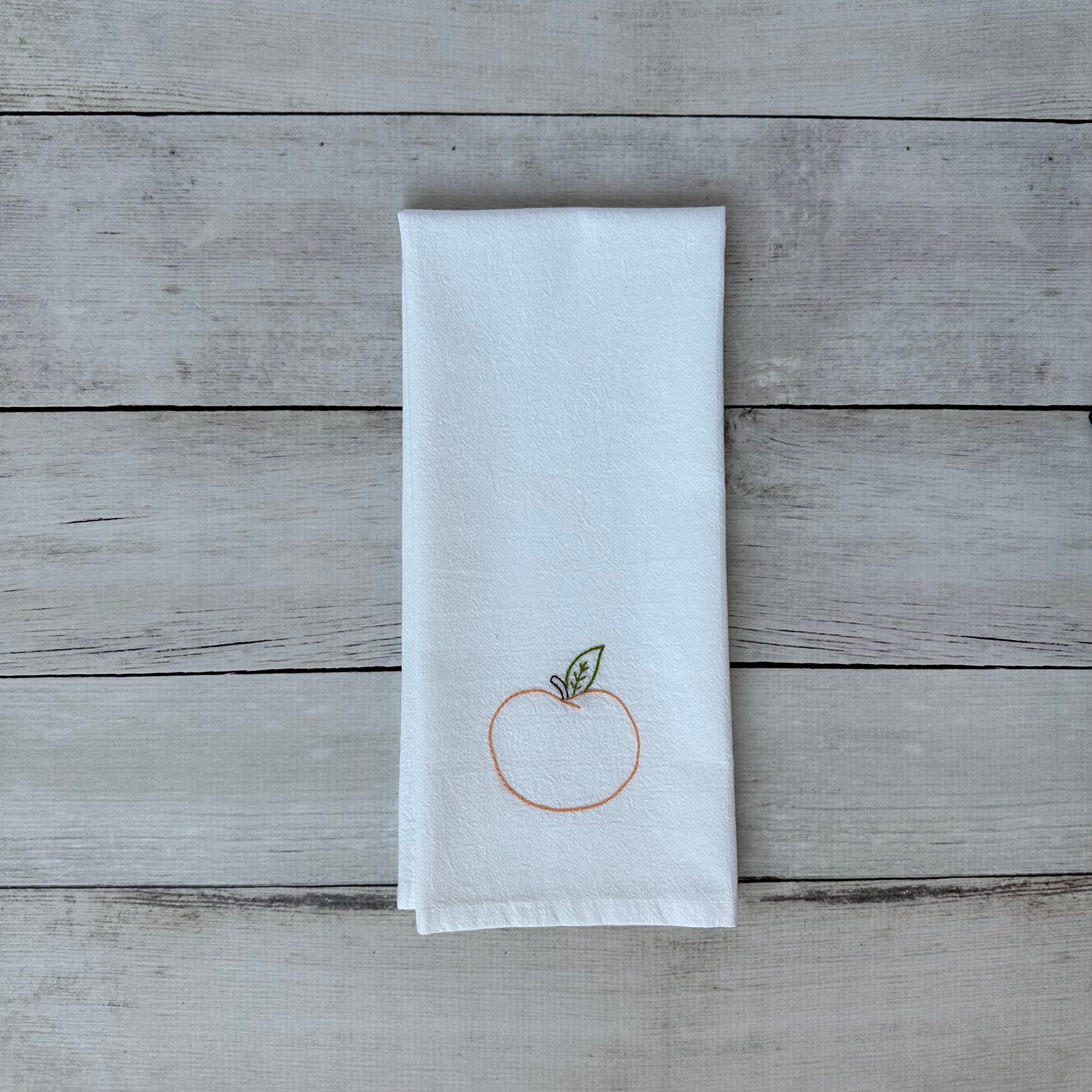 White tea towel with embroidered peach design on a wooden surface