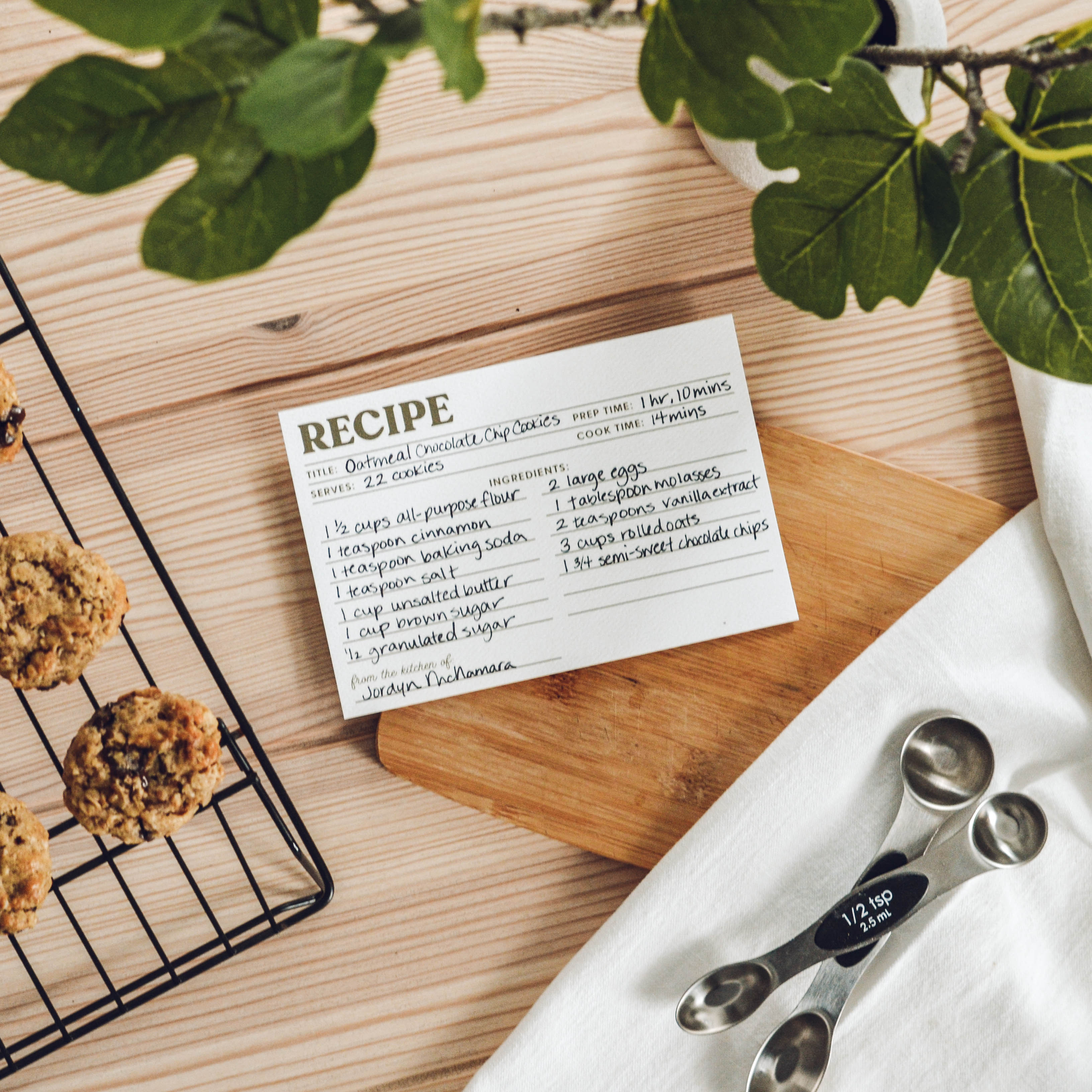 Recipe card for oatmeal raisin cookies on a wooden board with cookies and measuring spoons.