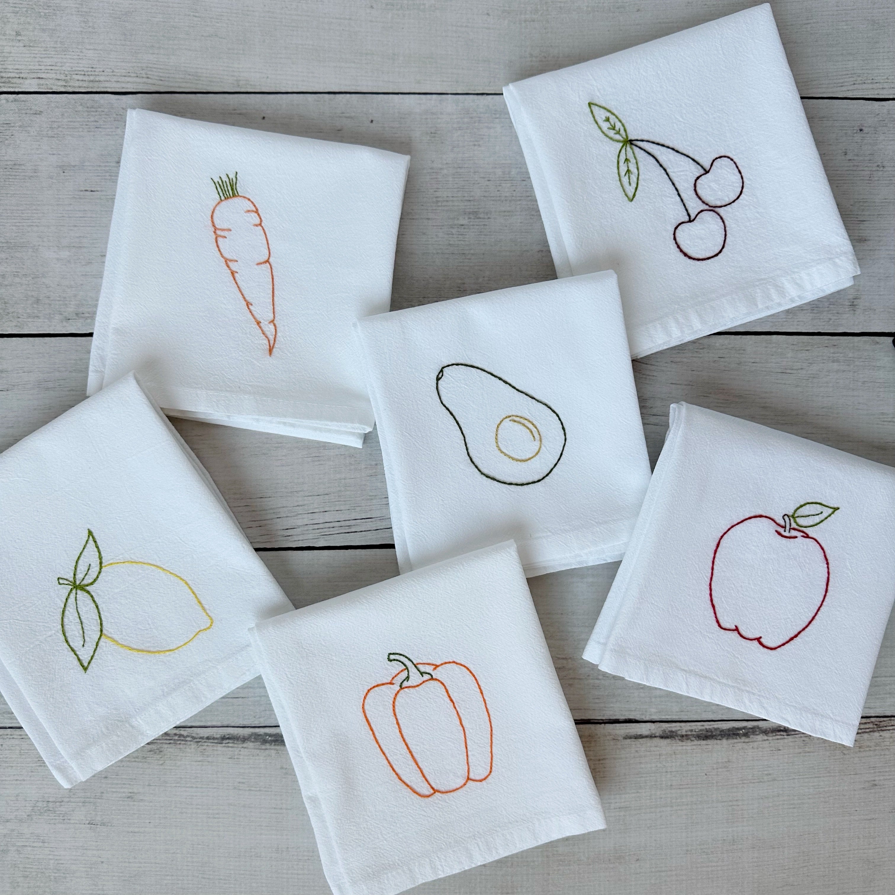 Set of tea towels with embroidered fruit and vegetable designs on a wooden surface