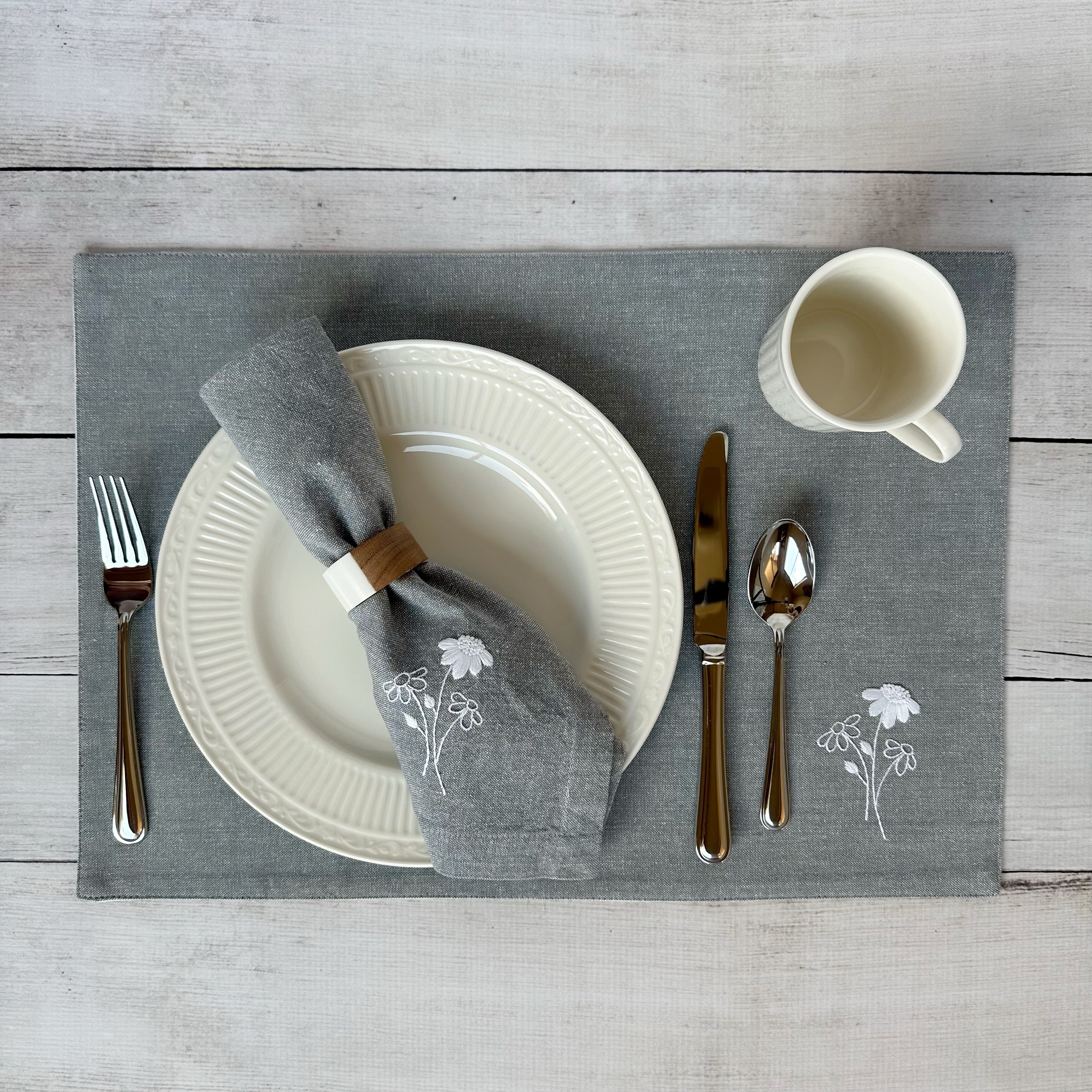 Dining table setting with white plates, silverware, and embroidered cotton napkin and placemat on a wooden surface.