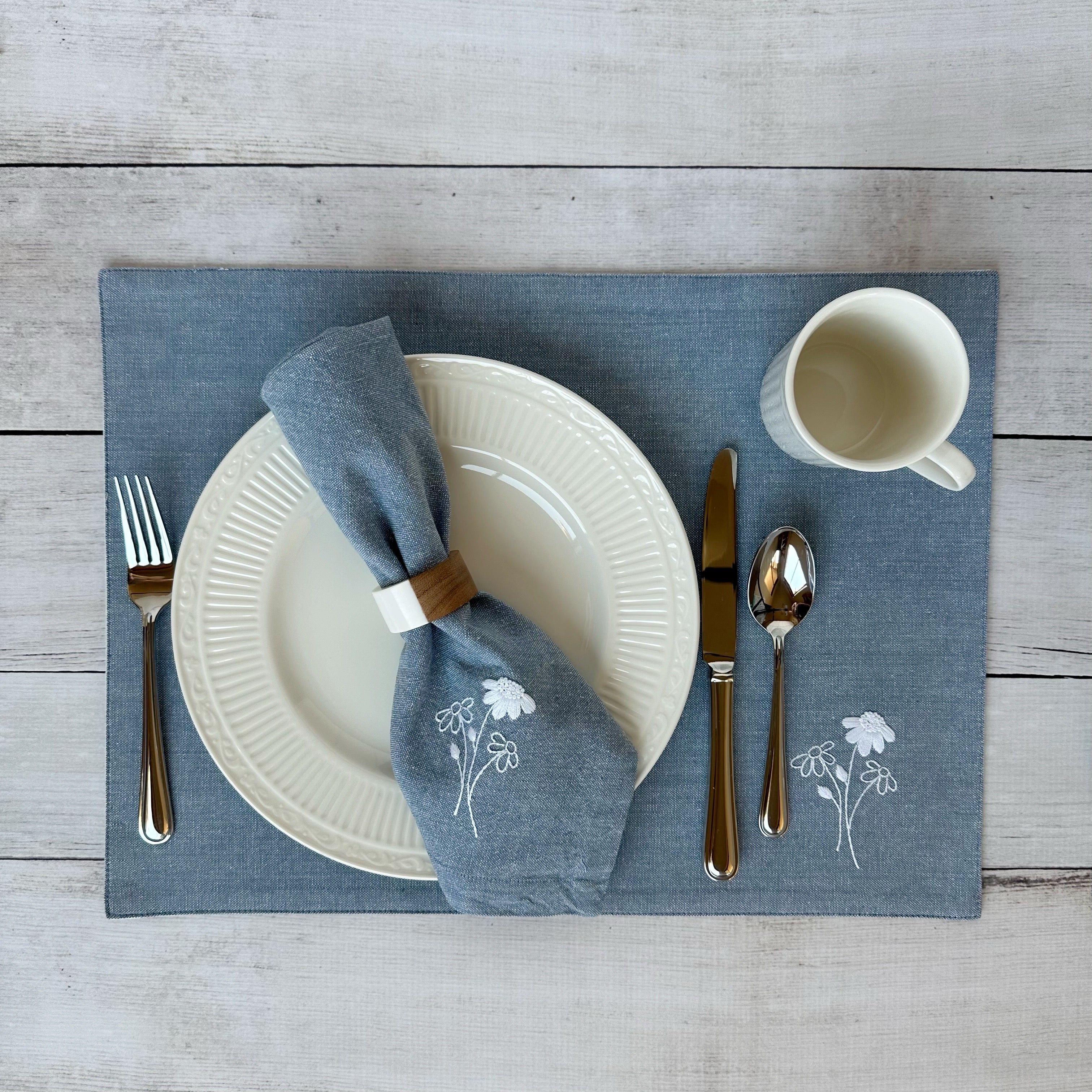Dining table setting with white plates, silverware, and embroidered cotton napkin and placemat on a wooden surface.