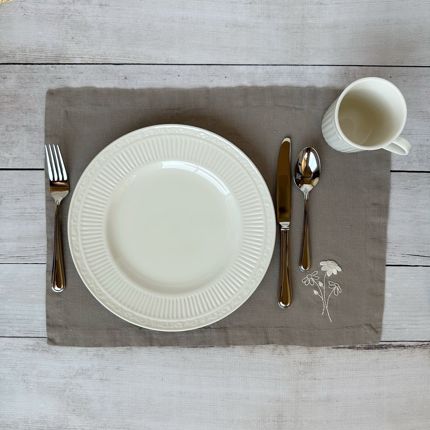 White china plate with silverware on a taupe placemat with embroidered flowers on a wooden surface