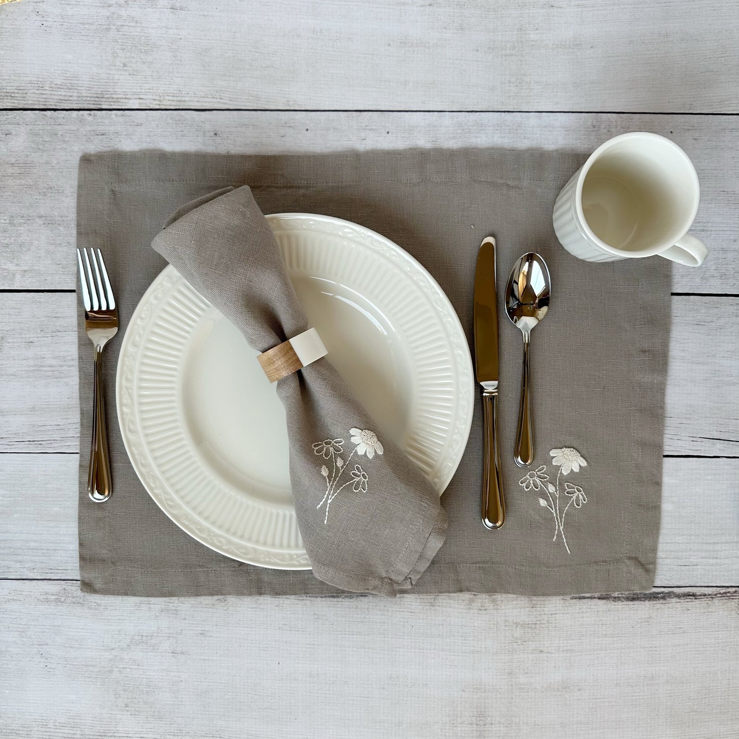 Dining table setting with white plates, silverware, and embroidered linen napkin and placemat on a wooden surface.