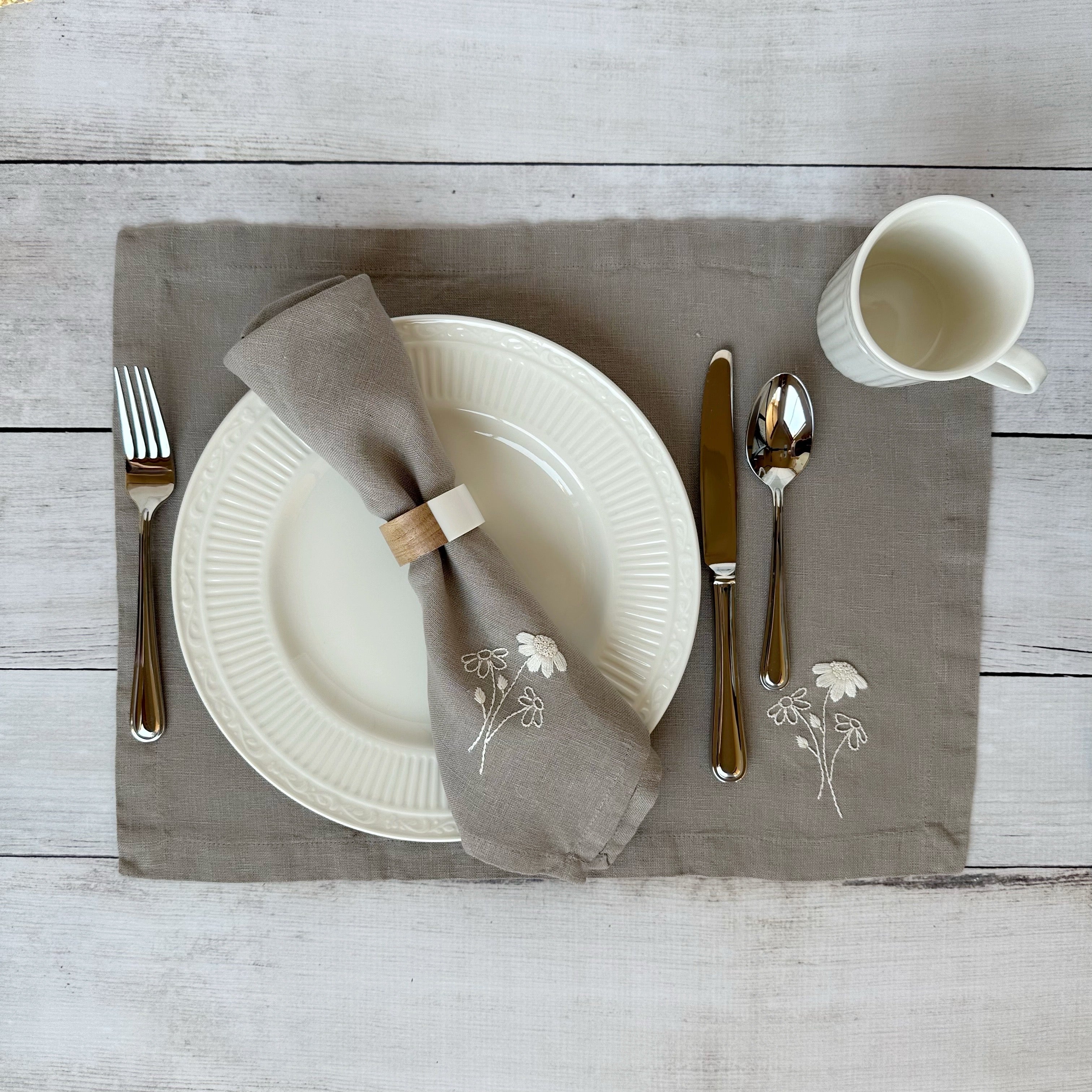Dining table setting with white plates, silverware, and embroidered linen napkin and placemat on a wooden surface.