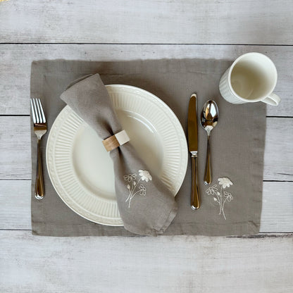 Dining table setting with white plates, silverware, and embroidered linen napkin and placemat on a wooden surface.