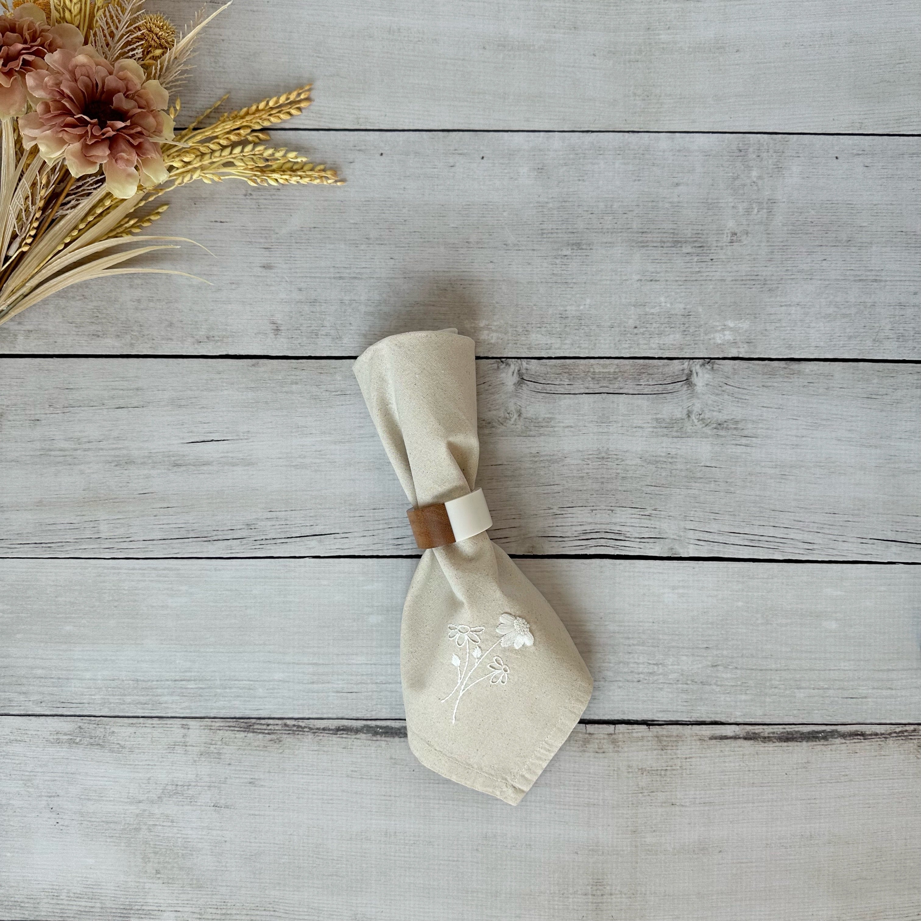 Beige cotton napkin with floral embroidery on a wooden surface with flowers.