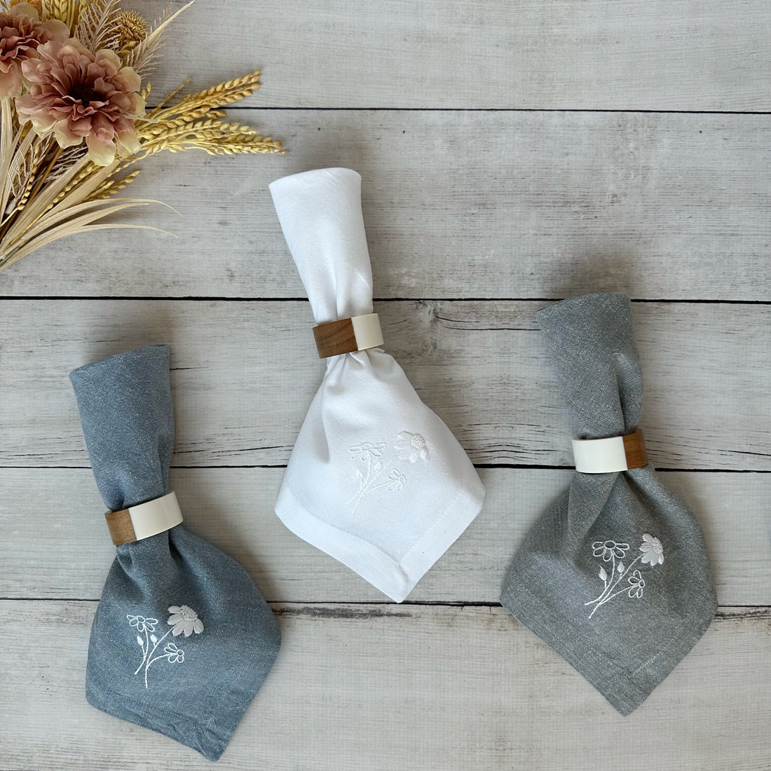 Gray, blue and white cotton napkins with floral embroidery on a wooden surface with flowers.