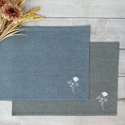 Two placemats with floral embroidery on a wooden surface.