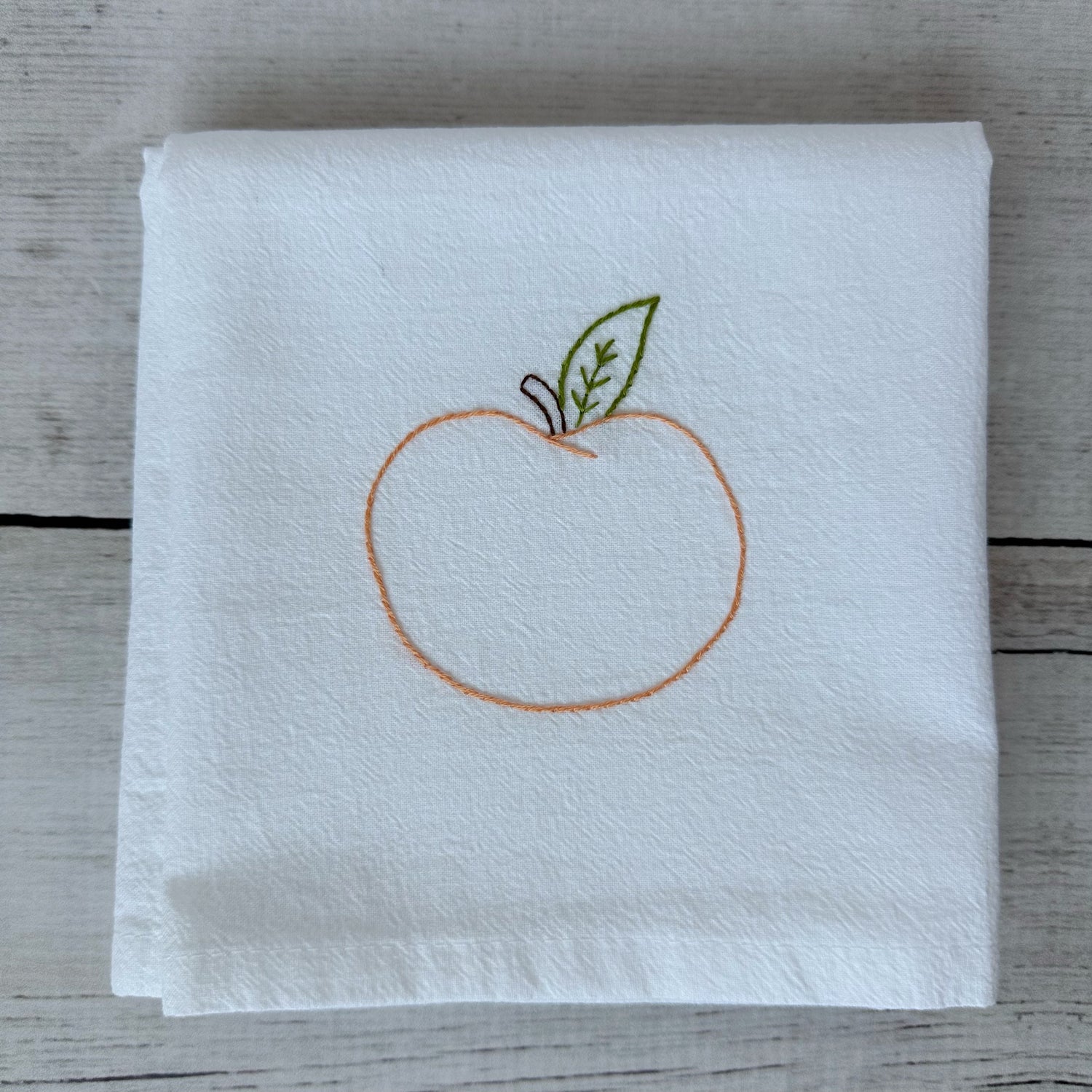 White tea towel with embroidered peach design on a wooden surface