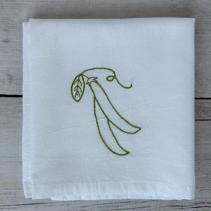 White tea towel with embroidered snap peas design on a wooden surface