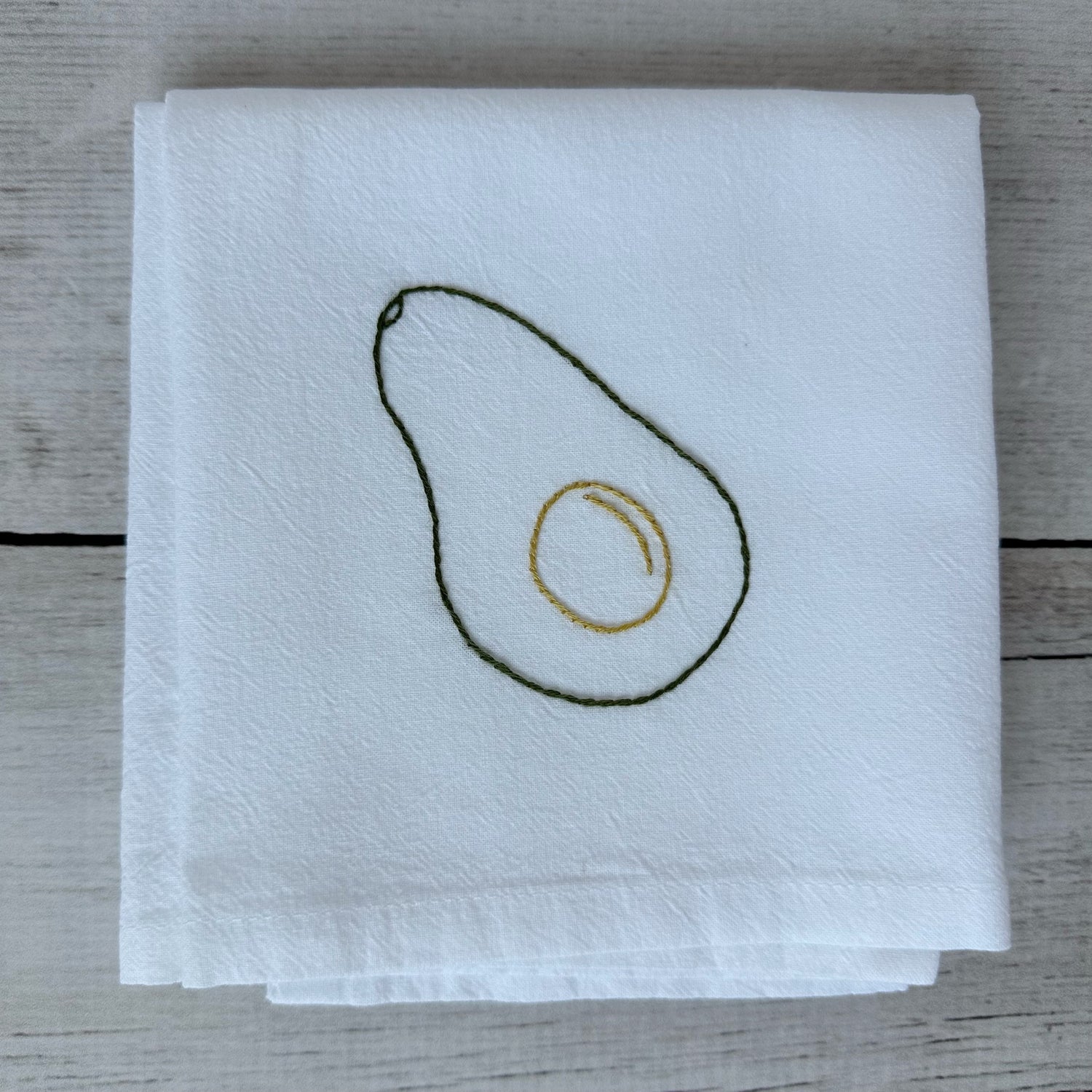 White tea towel with embroidered avocado design on a wooden surface