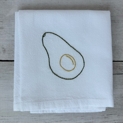 White tea towel with embroidered avocado design on a wooden surface