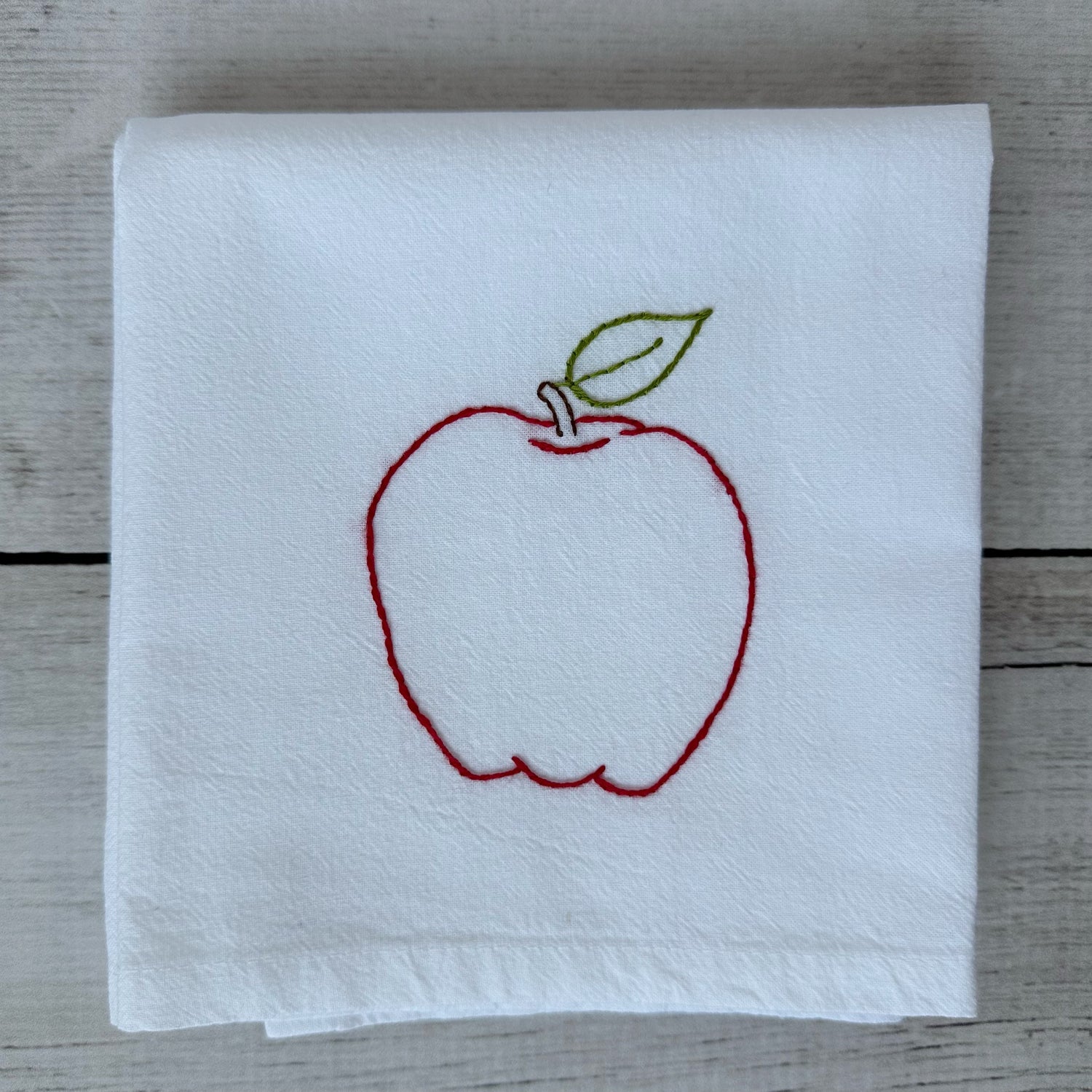 White tea towel with red embroidered apple design on a wooden surface