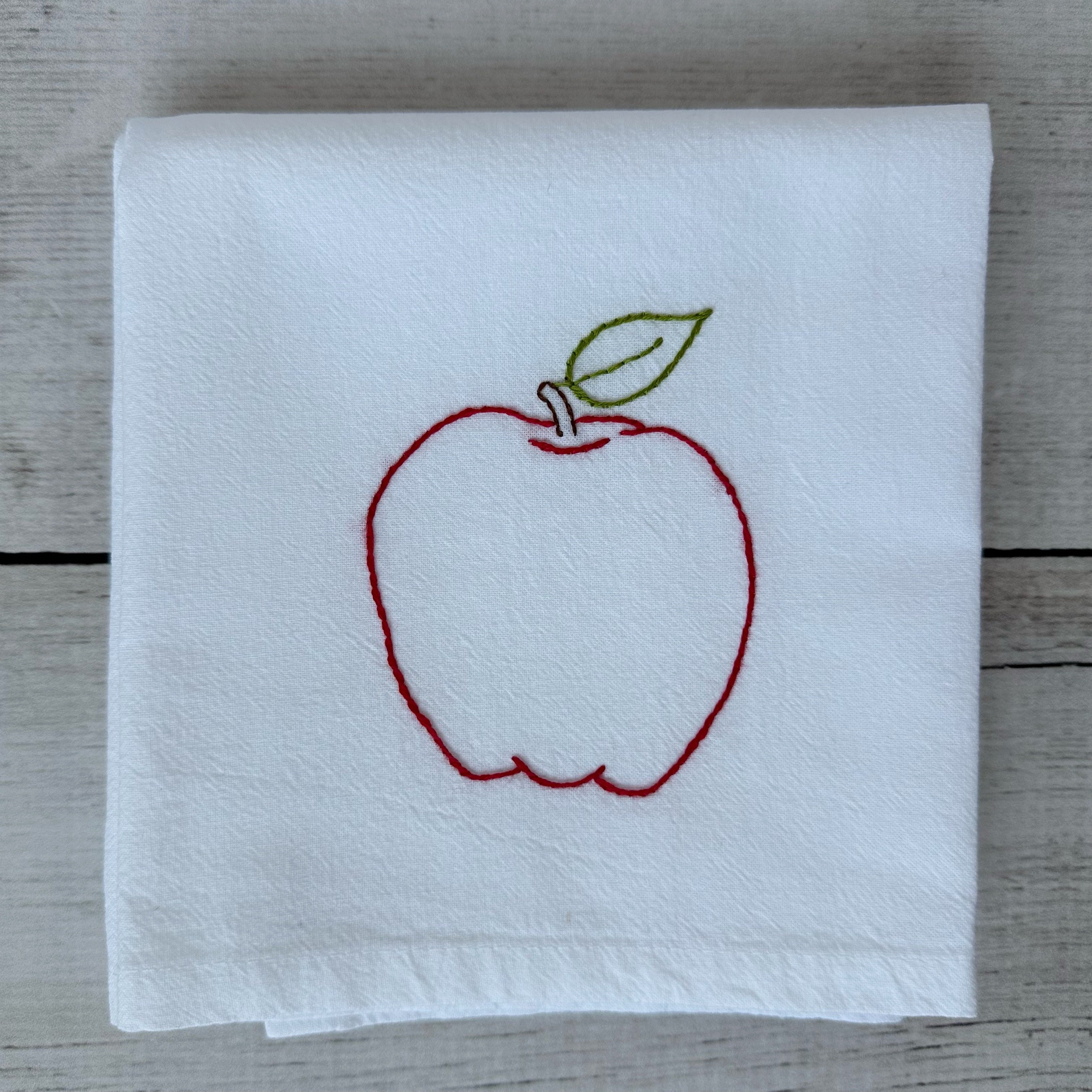 White tea towel with red embroidered apple design on a wooden surface